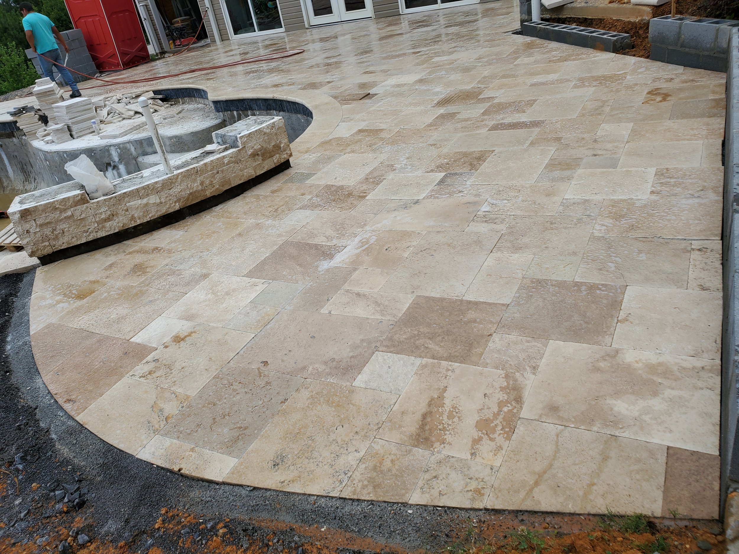Newly installed paver patio with beige and tan tiles in front of a house, with construction materials and workers present.