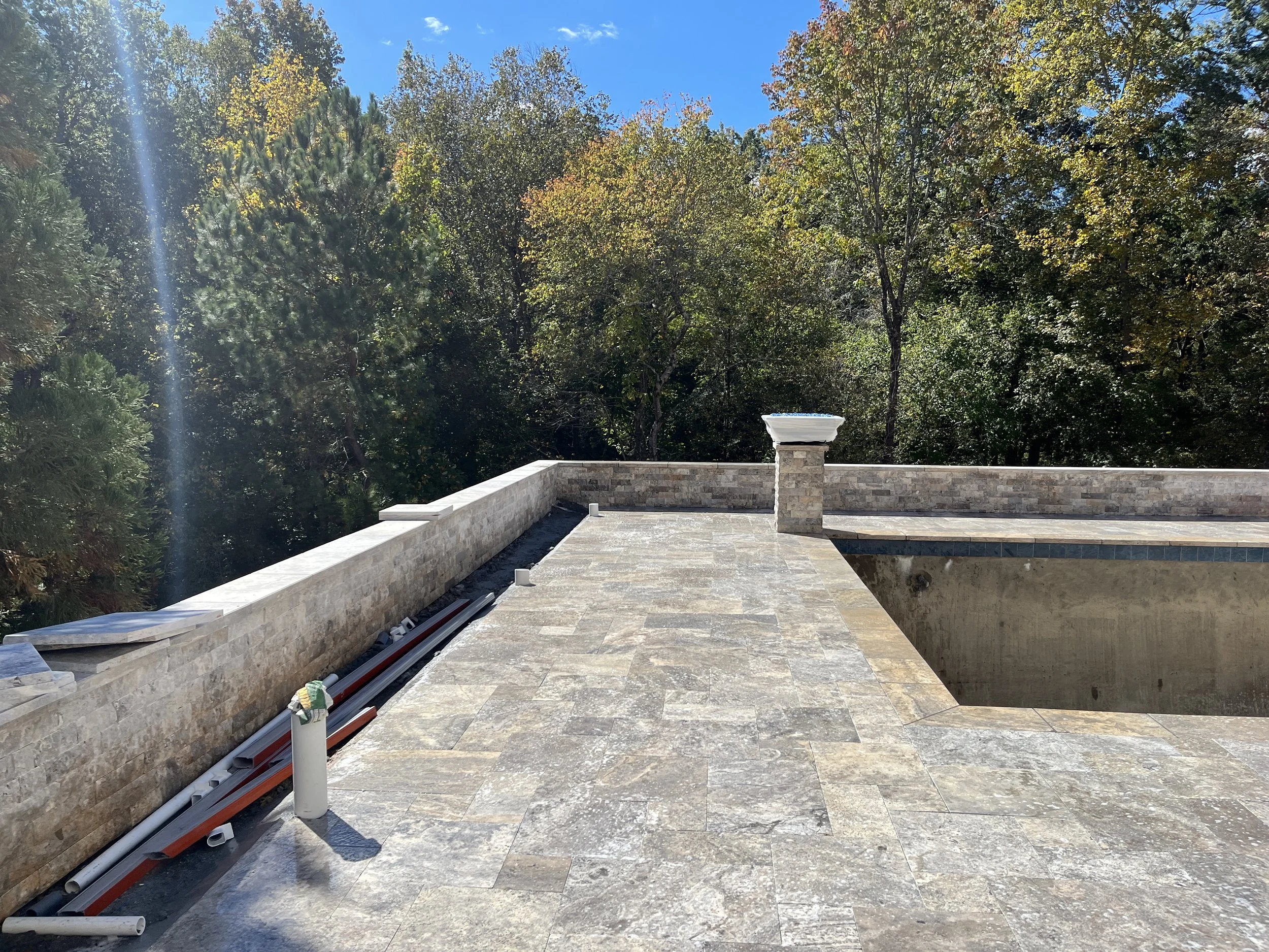 A backyard patio with stone tile flooring, a stone wall, and a hot tub under construction. There are several pipes and construction materials on the ground, and a small white structure, possibly a vent or pipe, protrudes from the wall. The background