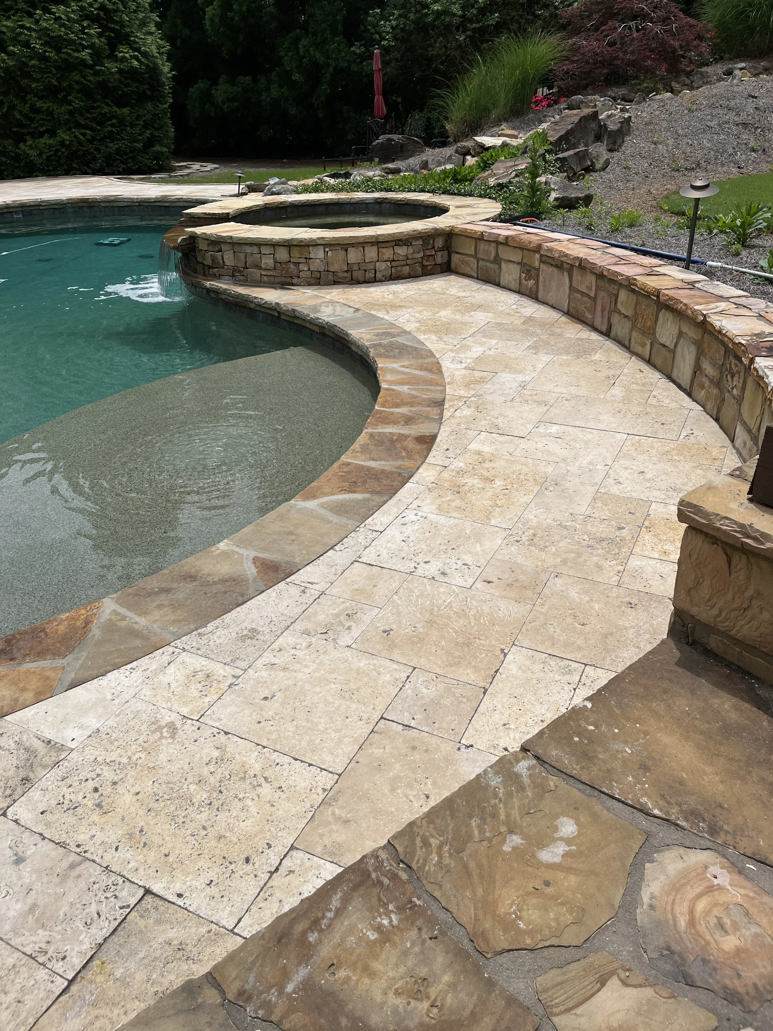 Backyard pool area with a stone deck, built-in hot tub, and landscaped garden with rocks and greenery.
