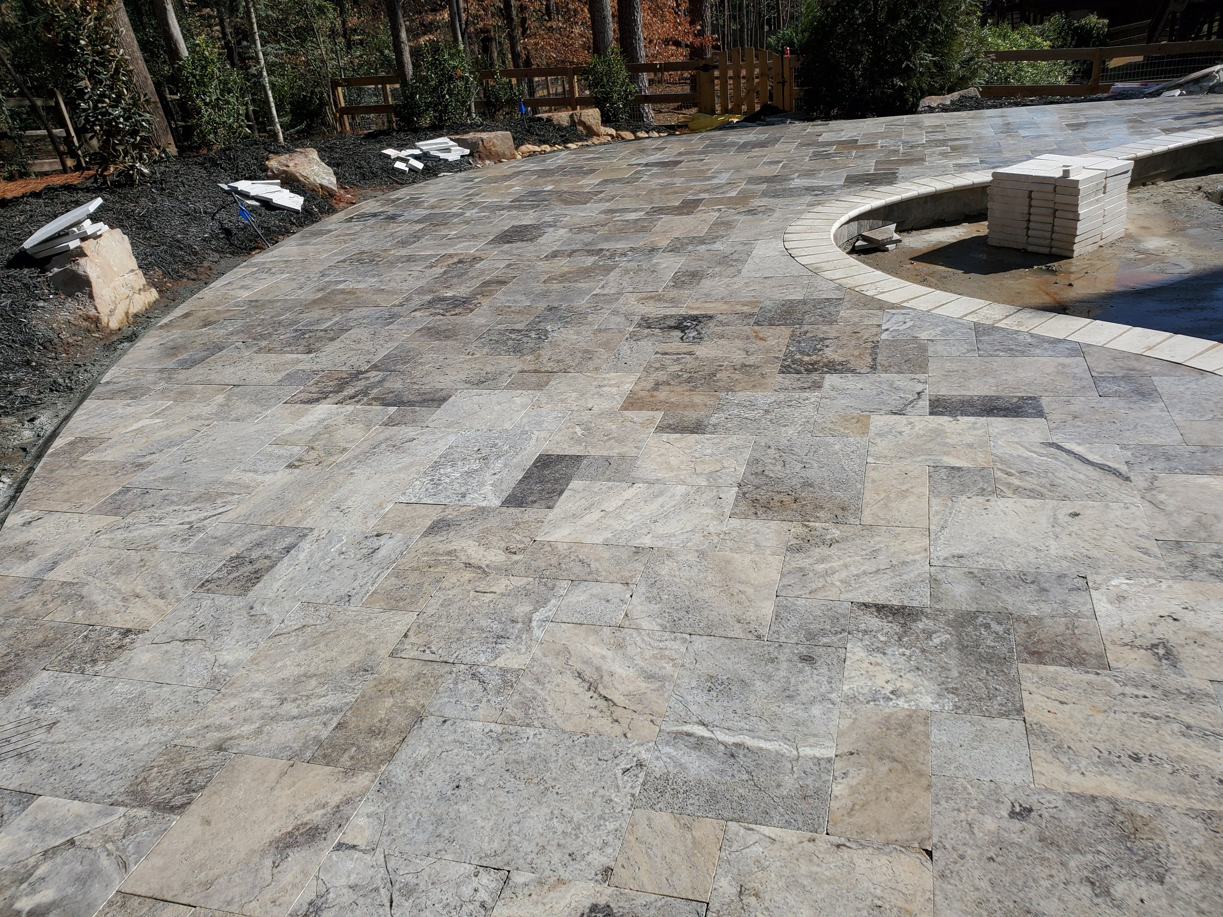 A newly paved outdoor patio made of rectangular stone tiles in various shades of gray and beige. There are some construction materials and tools on the ground, including white bricks stacked in a pile, scattered tiles, and some light fixtures. The ba