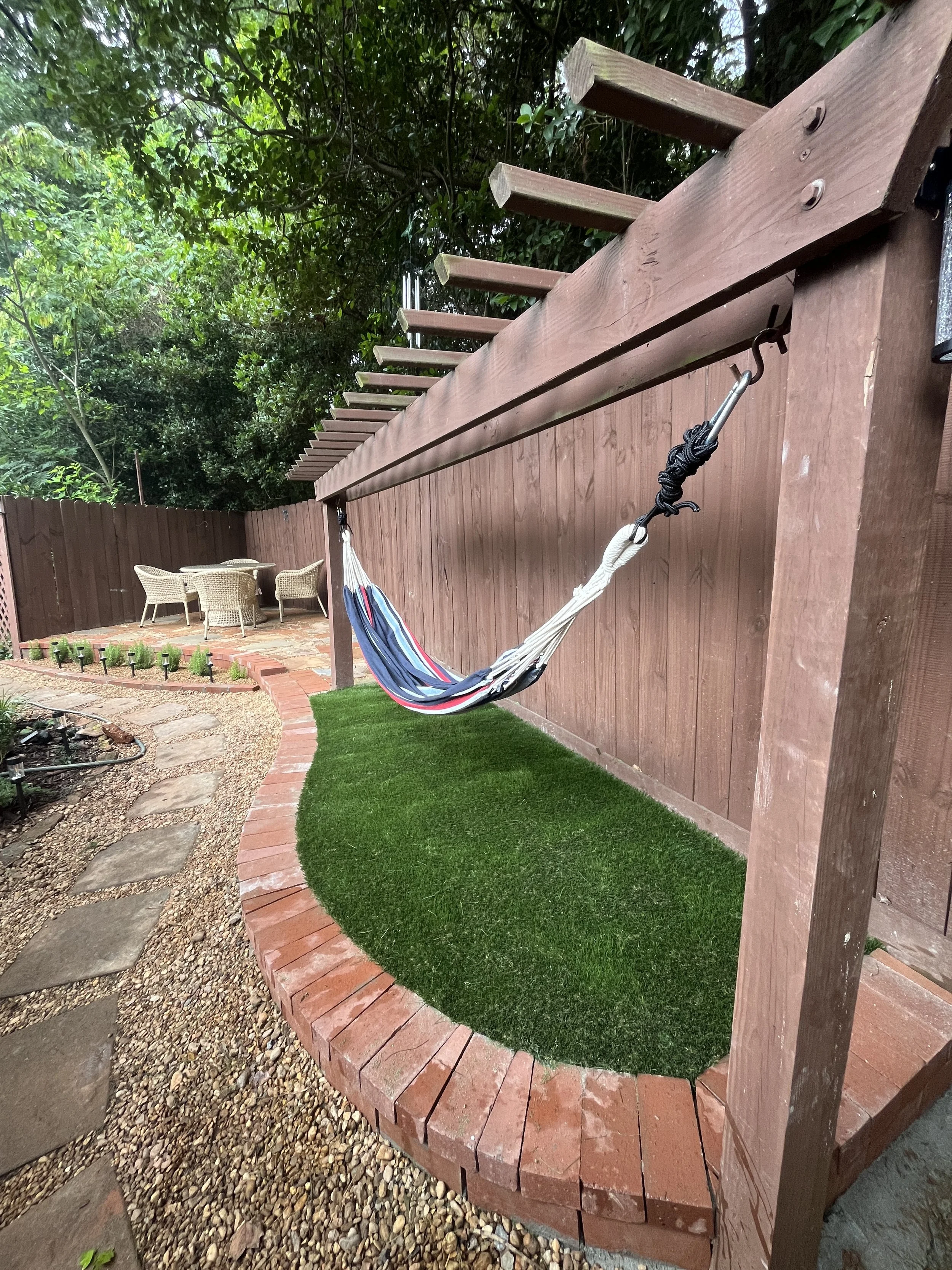 Backyard with a wooden structure that has a hammock hanging from it, a brick border, a small green grassy area, and a patio with wicker chairs, with trees and a wooden fence in the background.