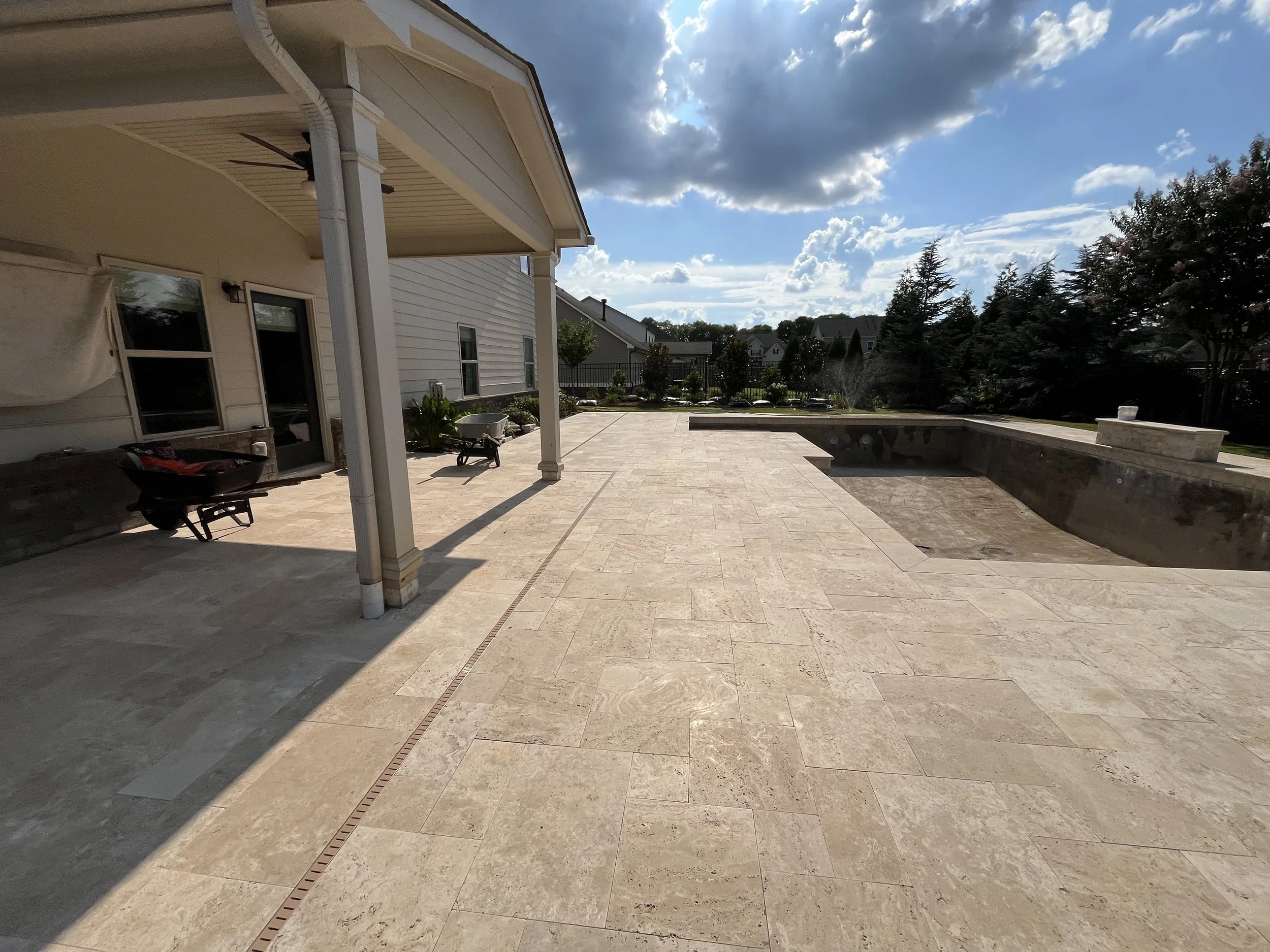 A spacious backyard patio area with beige tiled flooring, a covered section with an outdoor ceiling fan, and a low stone wall with a fountain. There are trees and residential houses in the background under a partly cloudy sky.