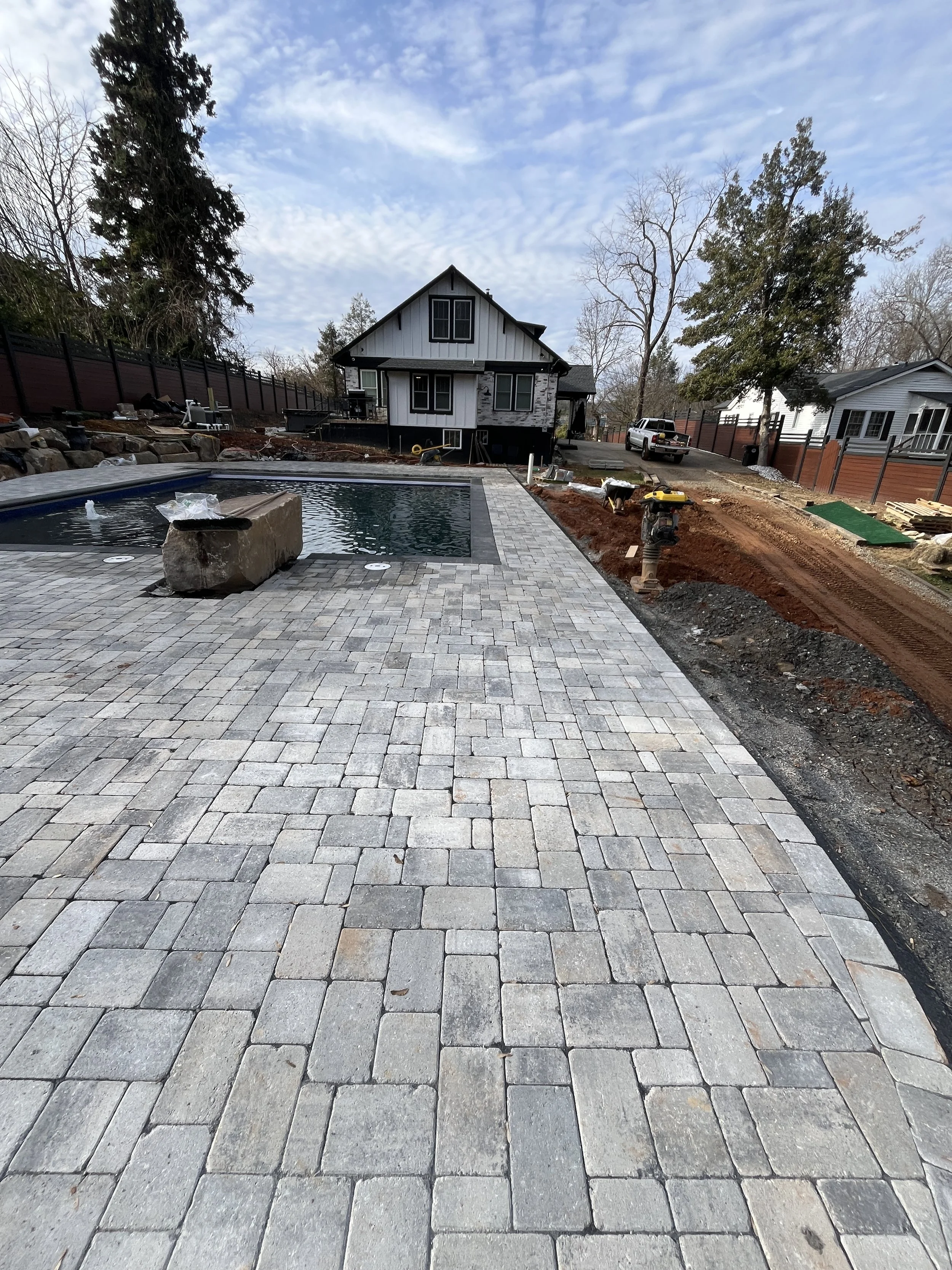 A backyard under construction with a paved stone patio and swimming pool in the foreground. A house with a dark roof and white siding, trees, and a driveway with a truck in the background. The site shows ongoing landscaping and construction work.