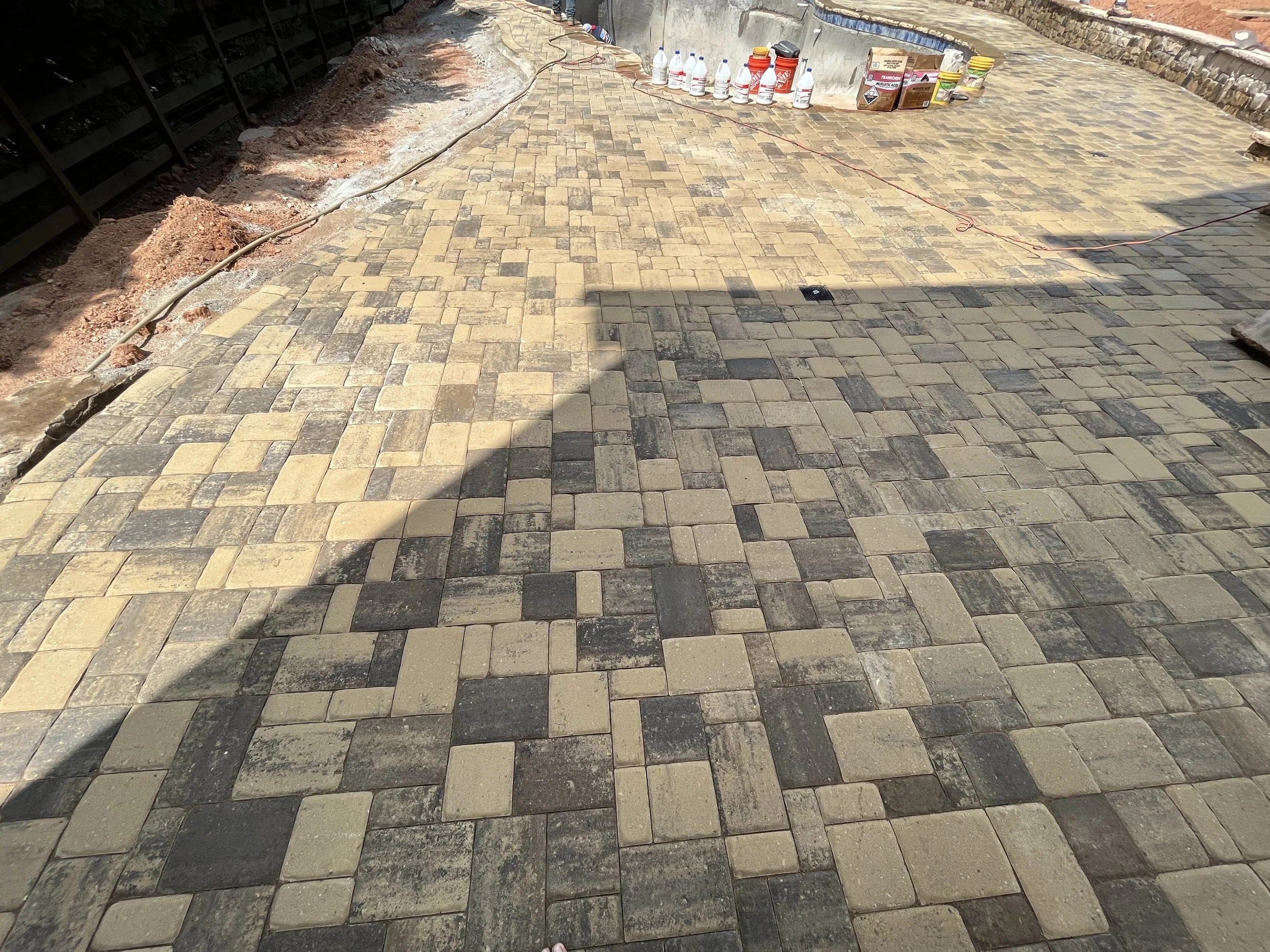 A paved outdoor pathway under construction with yellow and gray brick paving stones, construction supplies and tools along the side, and a shadow cast over part of the path.