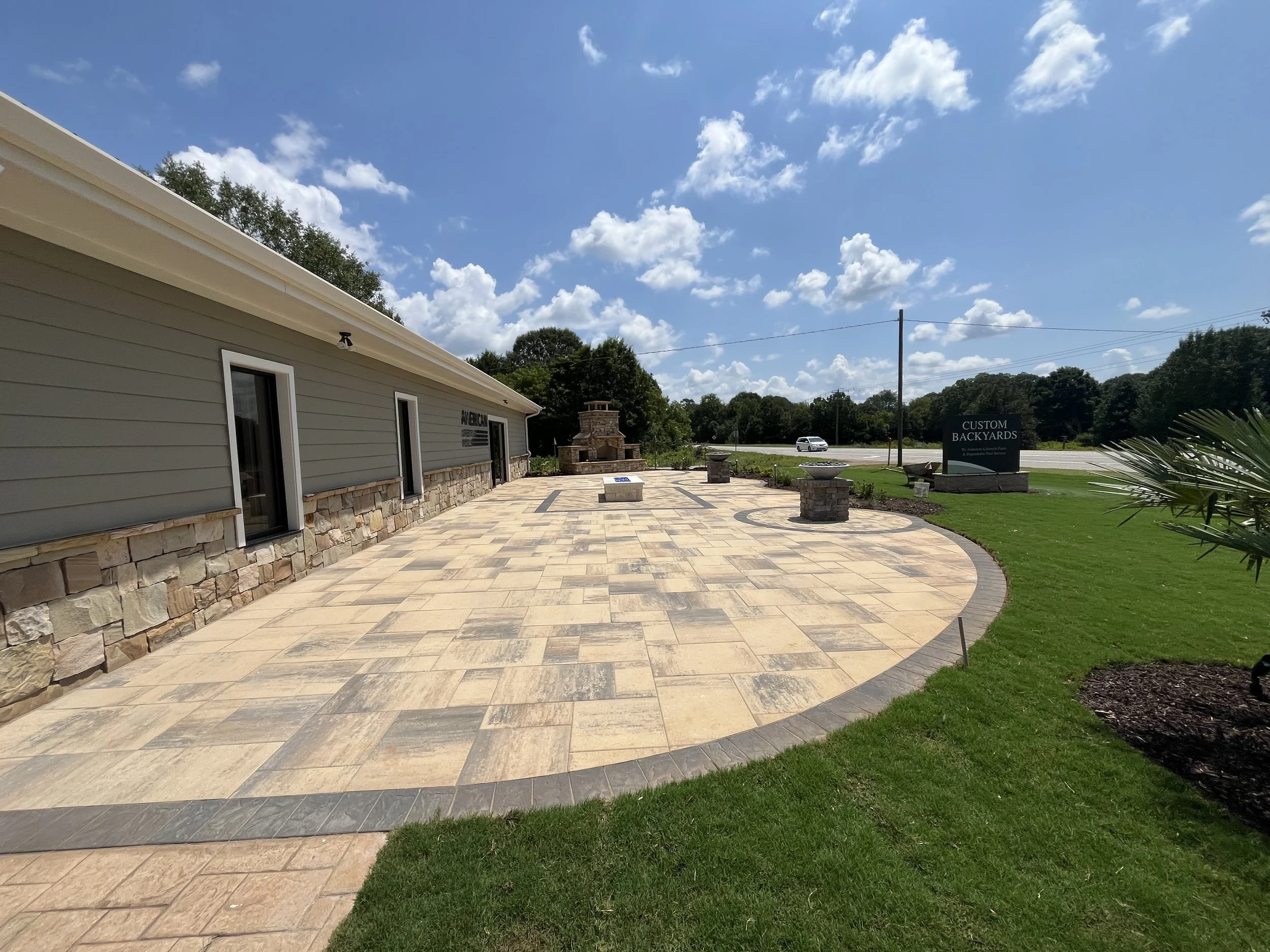 Exposed brick patio with a stone fireplace, gray siding building, well-manicured grass, sign that reads 'Custom Backyards', and a road with moving cars under a blue sky with puffy clouds.