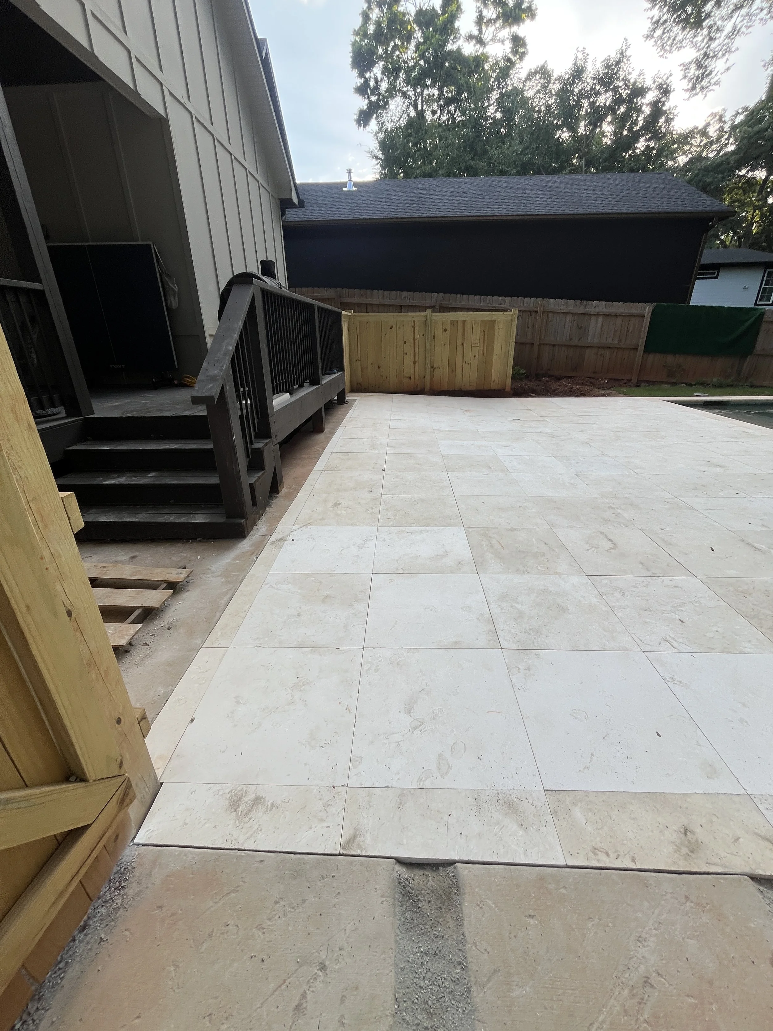 Backyard patio area with new light-colored stone tiles, a small step leading to a deck with black railing, a wooden fence, and trees in the background with a partly cloudy sky.