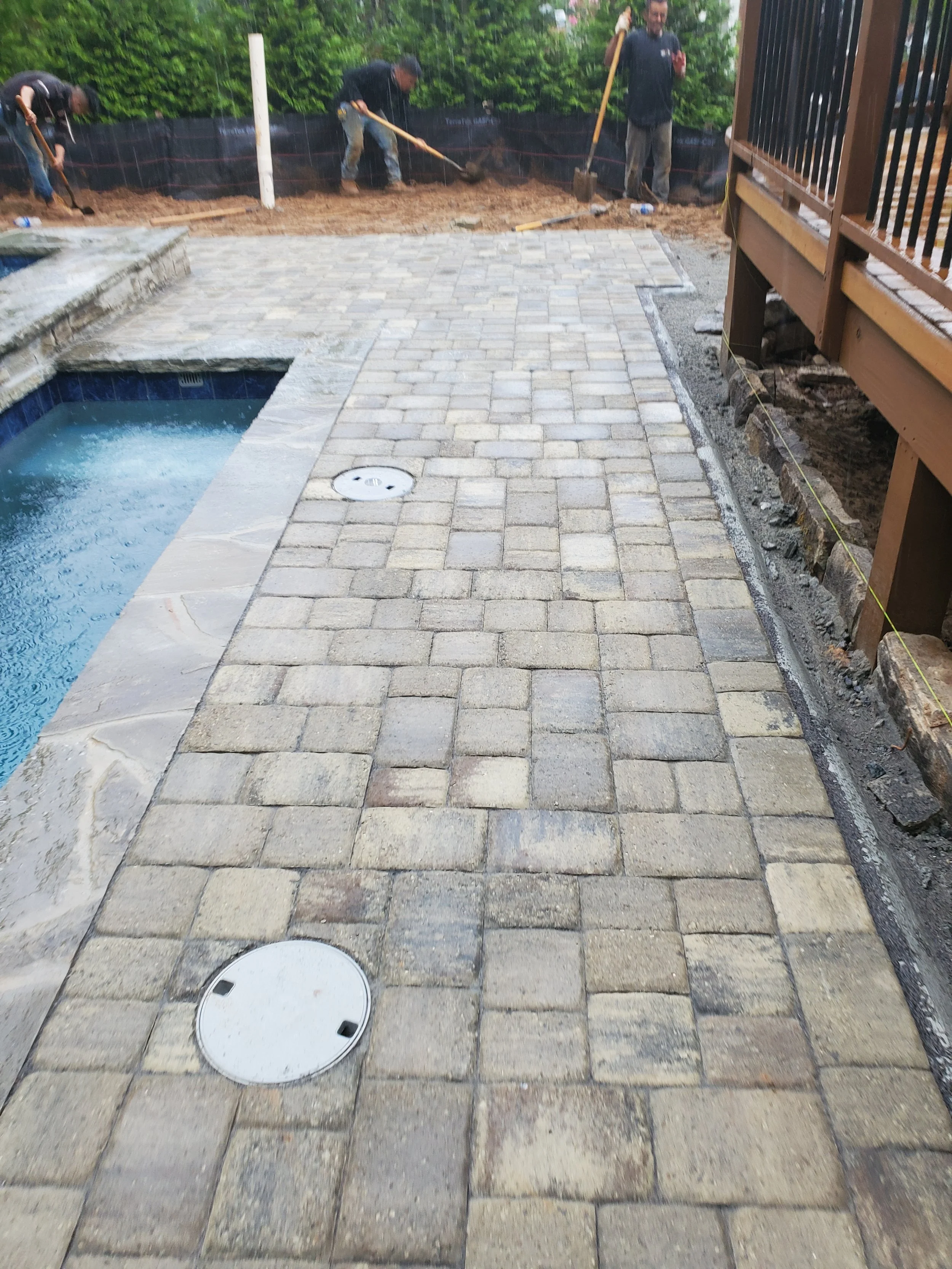 Three workers are constructing or repairing a paved backyard area. They are spreading dirt on the edge of the paved area. There is a swimming pool on the left and a wooden deck on the right. Construction materials and tools are scattered around.