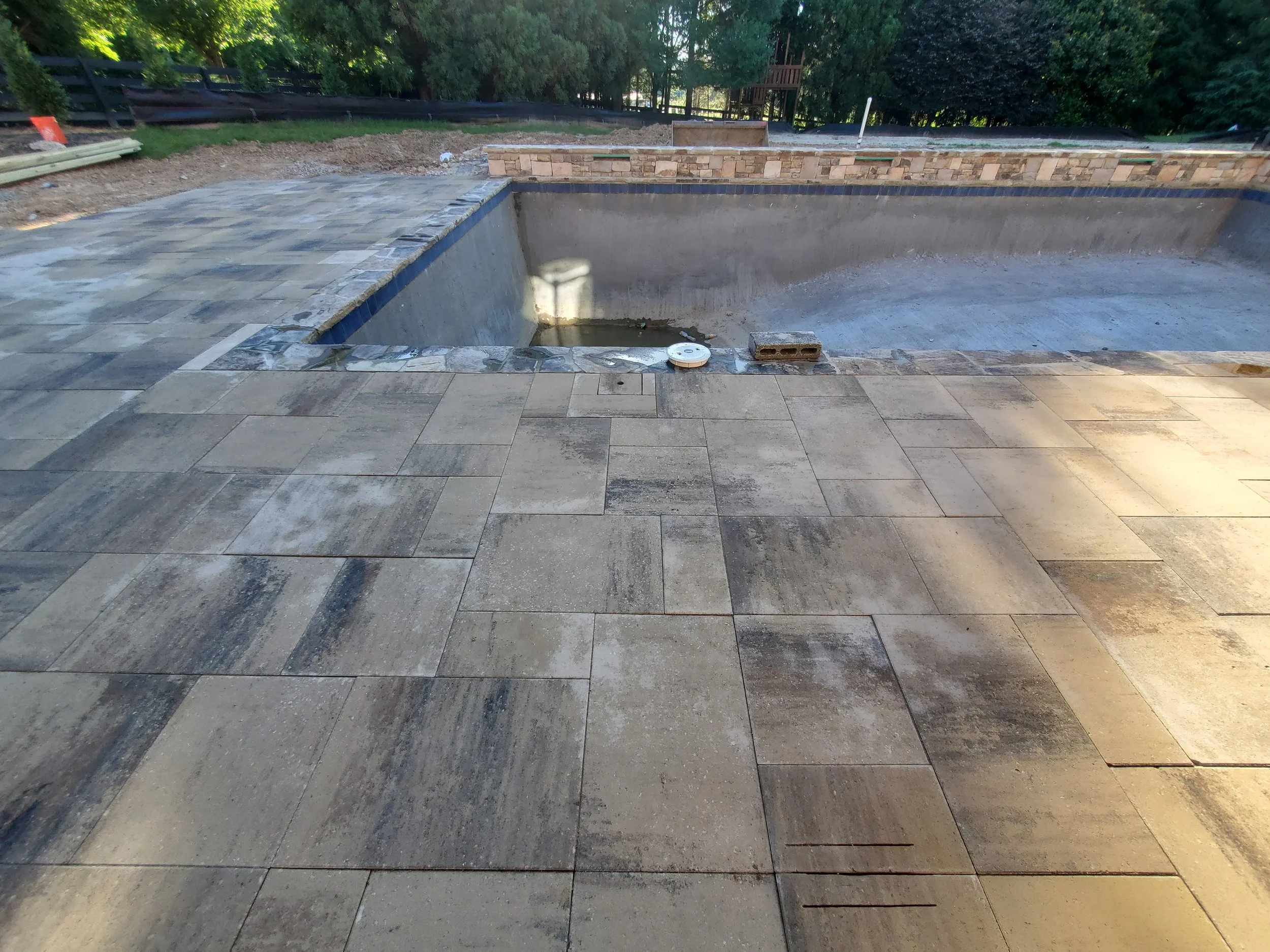 Empty swimming pool under construction with tiled deck surrounding it and construction materials nearby, surrounded by trees.
