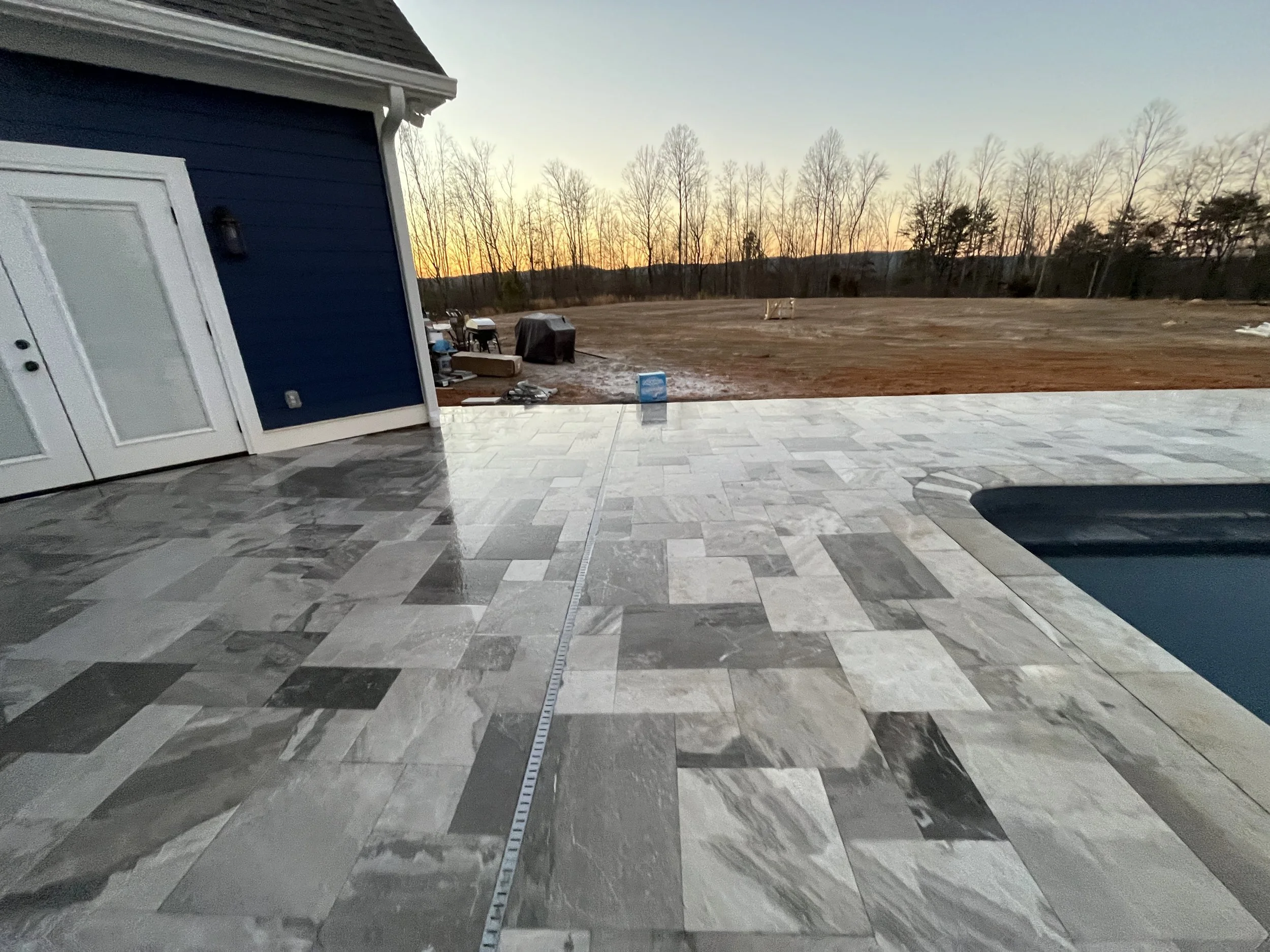 Back patio with tiled flooring, adjacent to a blue house with a door and light fixture. In the background, a large open dirt yard with some covered items, trees, and sunset sky.