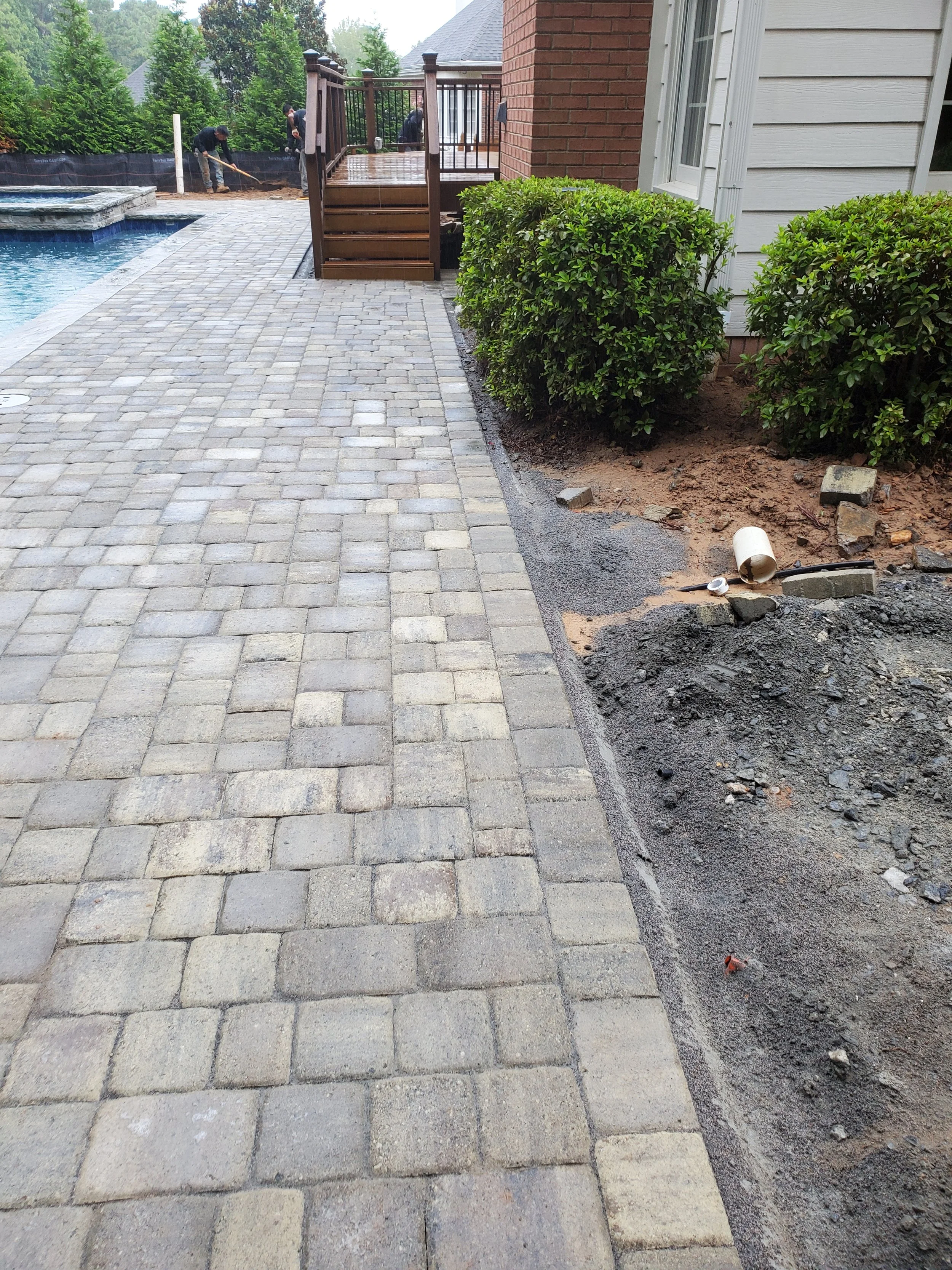 Backyard with brick house, gray wooden siding, a wooden deck with stairs, a swimming pool, and workers laying pavers or working on an outdoor project.