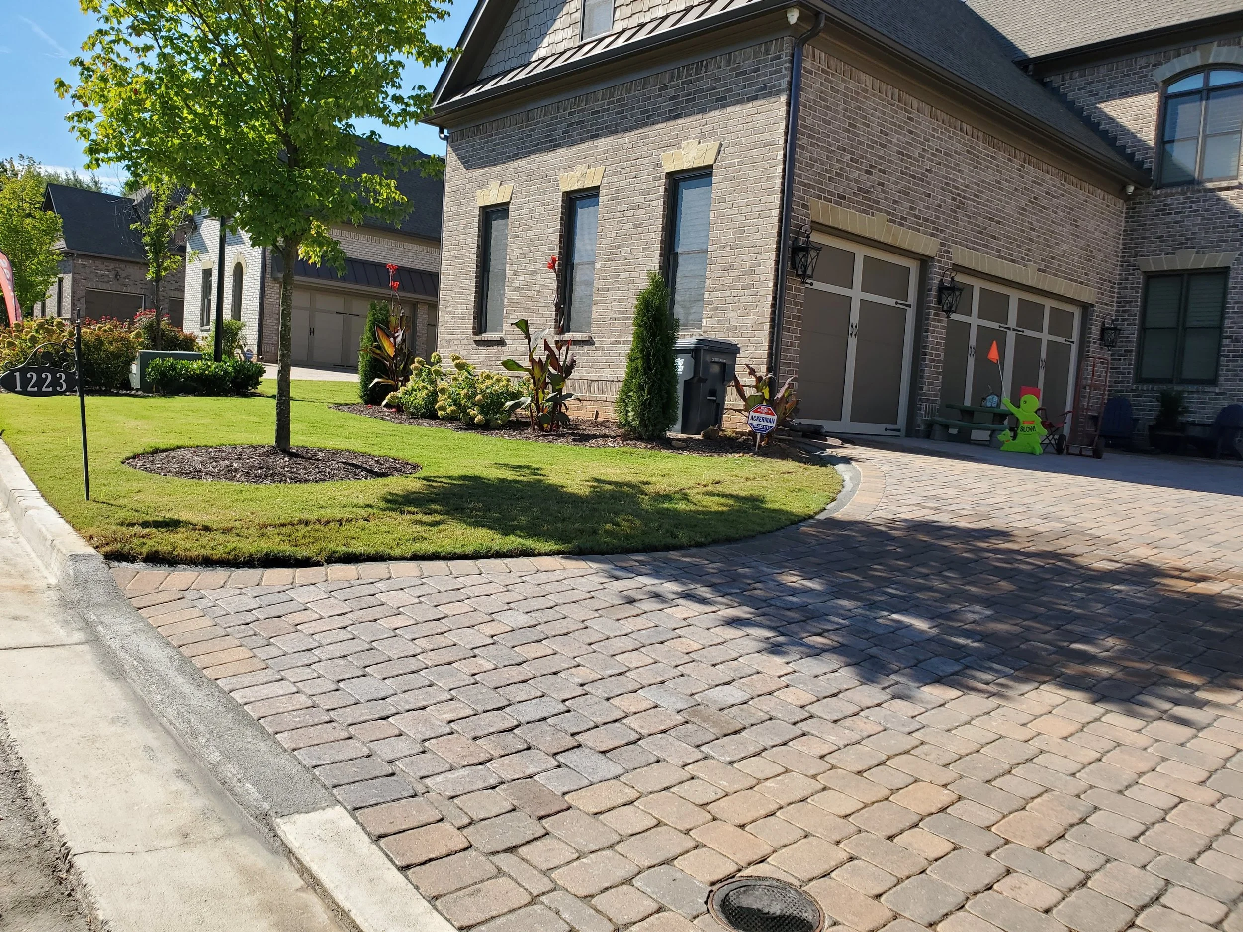A suburban house with a brick exterior, a gray garage door, and a well-maintained front yard with a tree, bushes, and flowers. There is a sidewalk, a curved driveway paved with bricks, and a lawn around the house. Decorations and a trash bin are near