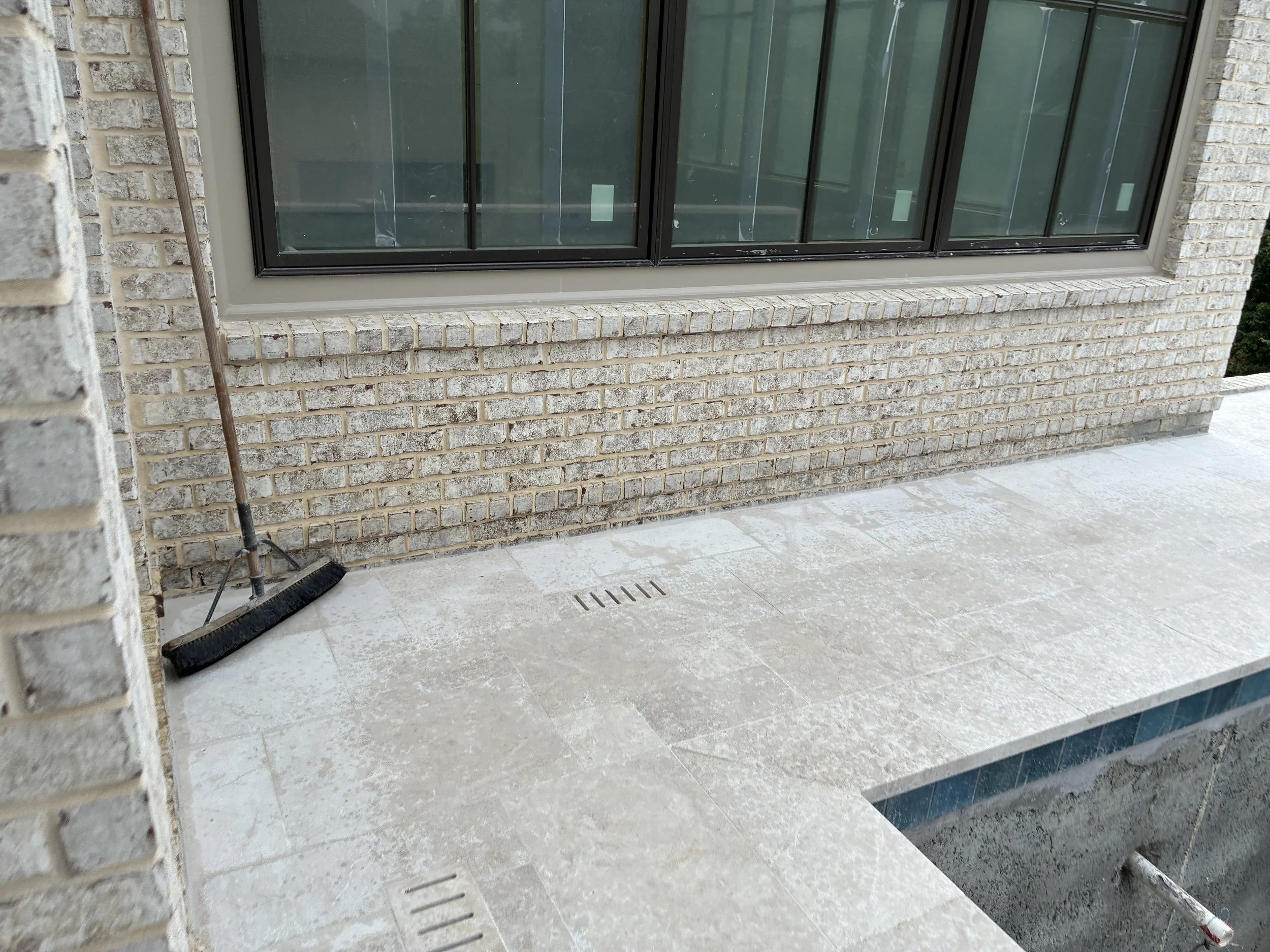 A brick exterior wall of a building with a large glass window. The window has black metal framing and is partially covered by yellow protective film. Below the window is a small ledge made of the same bricks. In the foreground, there is a shovel, and