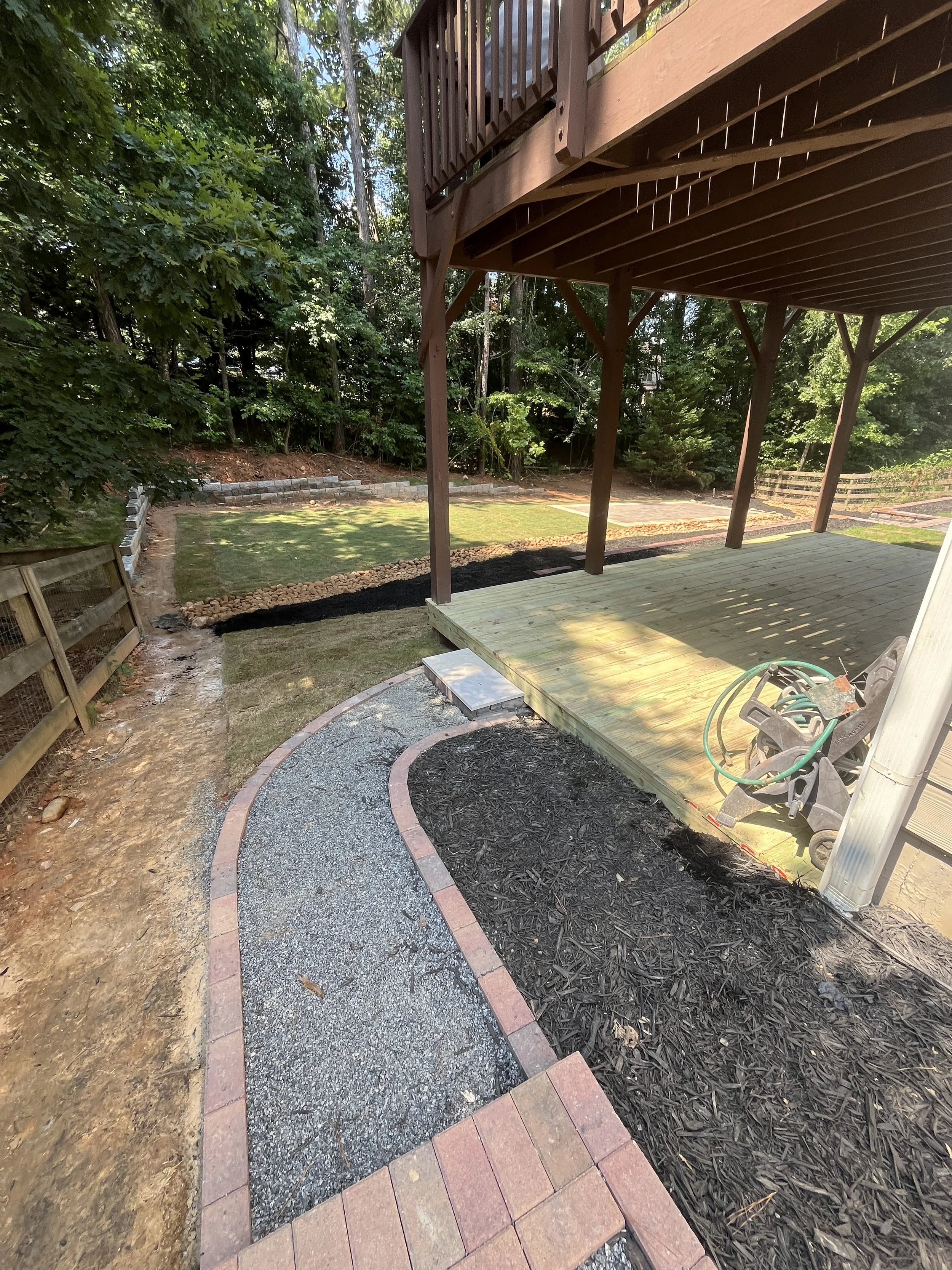 Backyard with a newly constructed wooden deck, garden bed with mulch, and a winding pathway with brick edging surrounded by grass and trees.