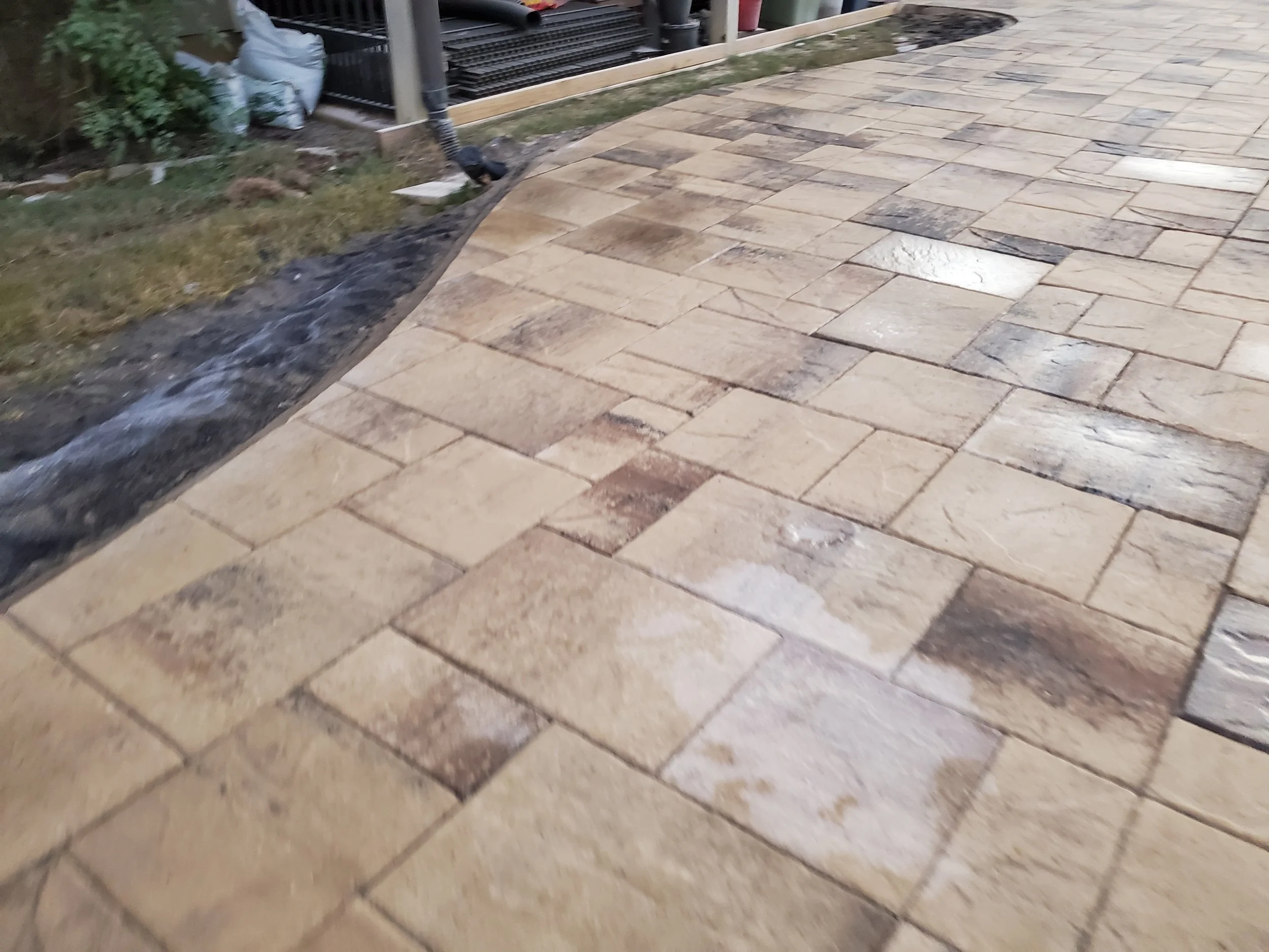 A newly installed stone patio with interlocking rectangular pavers in shades of tan, beige, and gray. There is a small patch of dirt and grass on the left side, with some green plants and a white bag nearby. In the background, there's construction or