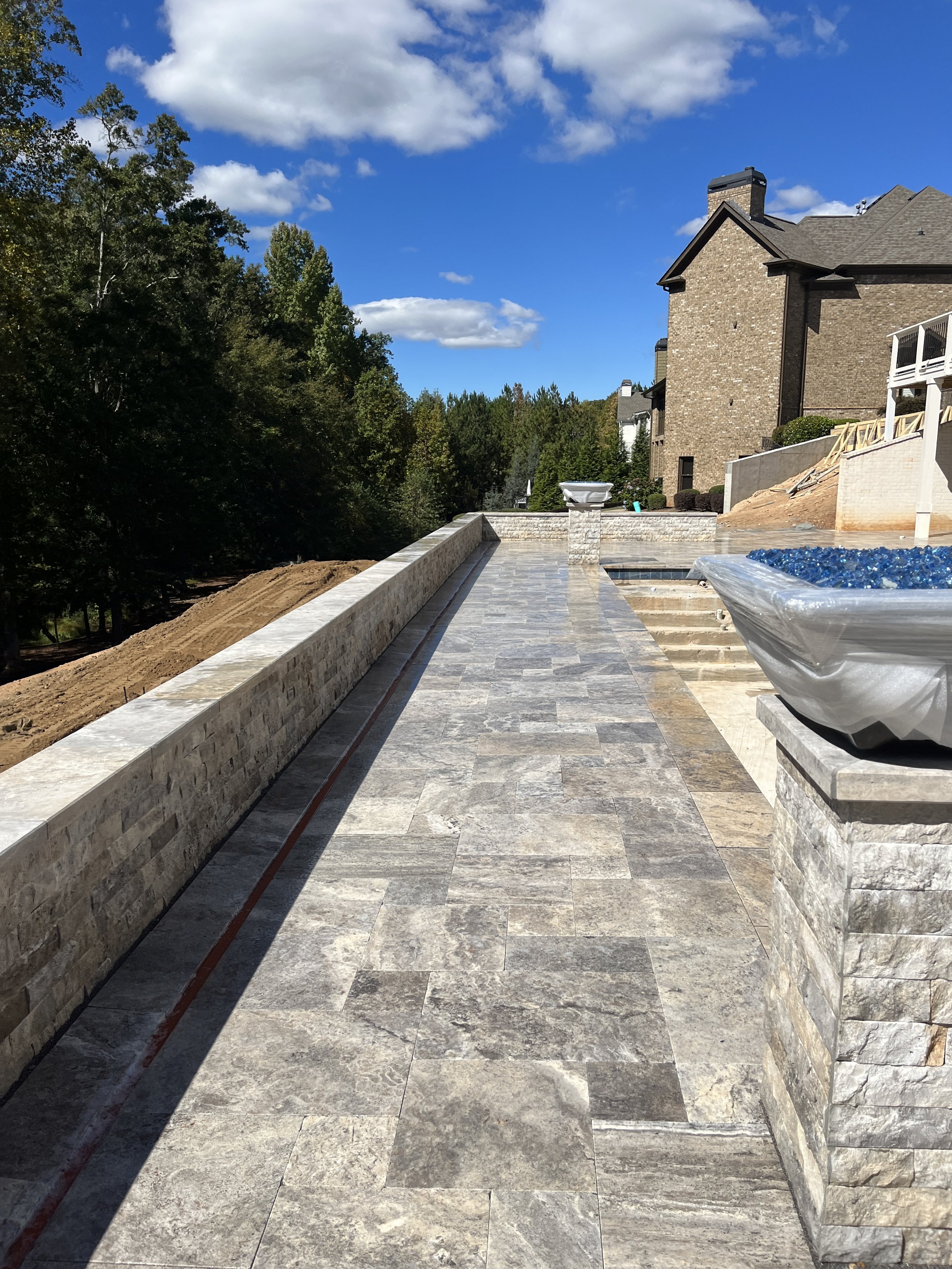Construction site with newly paved stone patio, brick wall, and partially built stairs, with a house, trees, and a blue sky with clouds in the background.