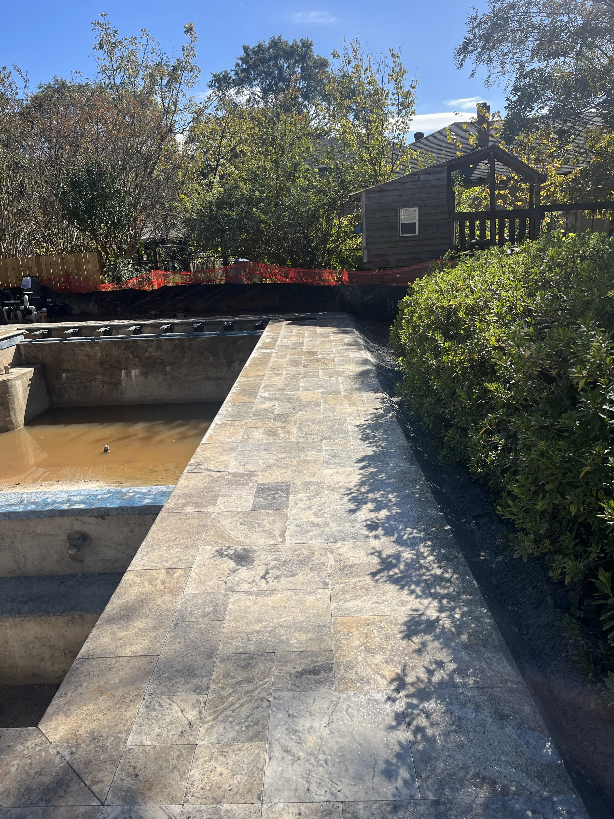 Side view of an outdoor construction site with a partially built hot tub, surrounding tiles, bushes, a wooden shed, trees, and a clear blue sky.