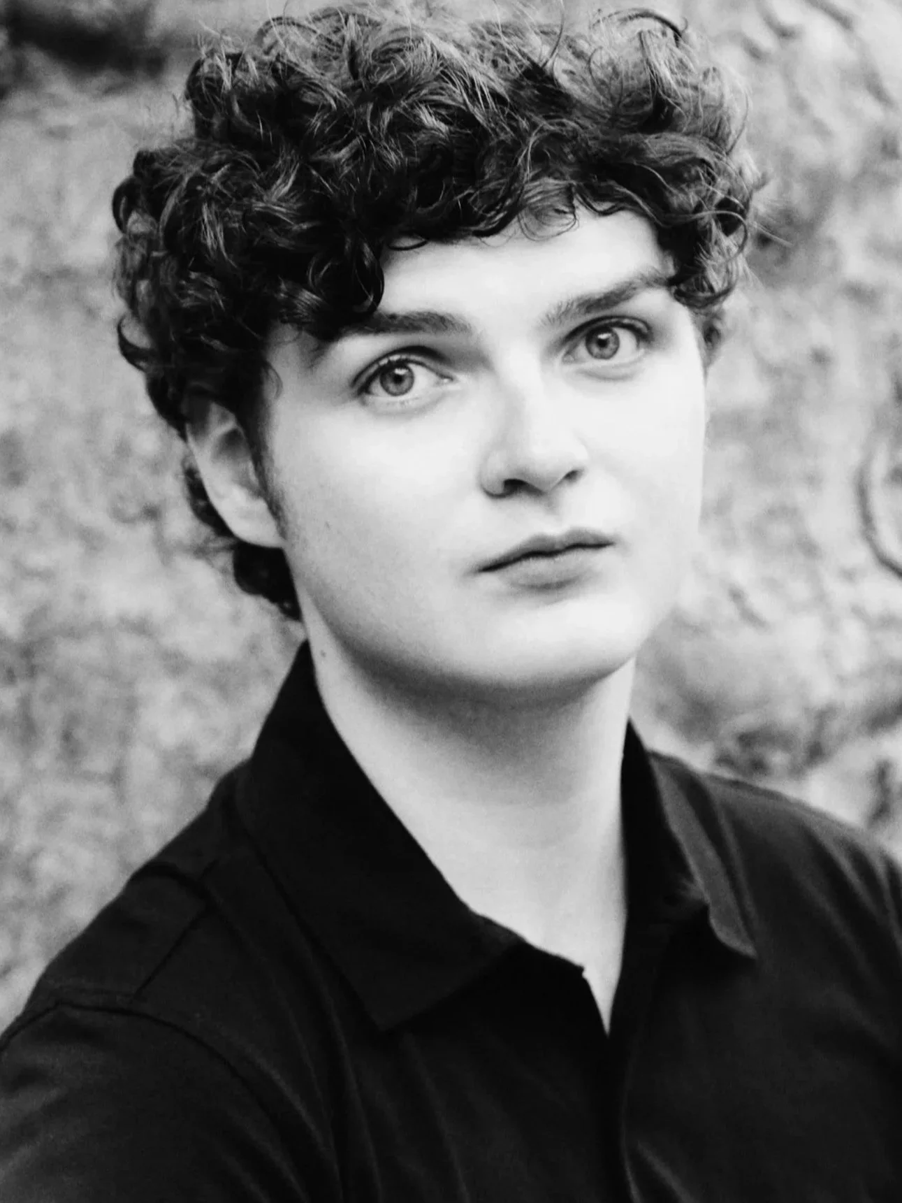Black and white portrait of a young man with short curly hair, light eyes, and wearing a dark collared shirt, outdoors, with a blurred background.