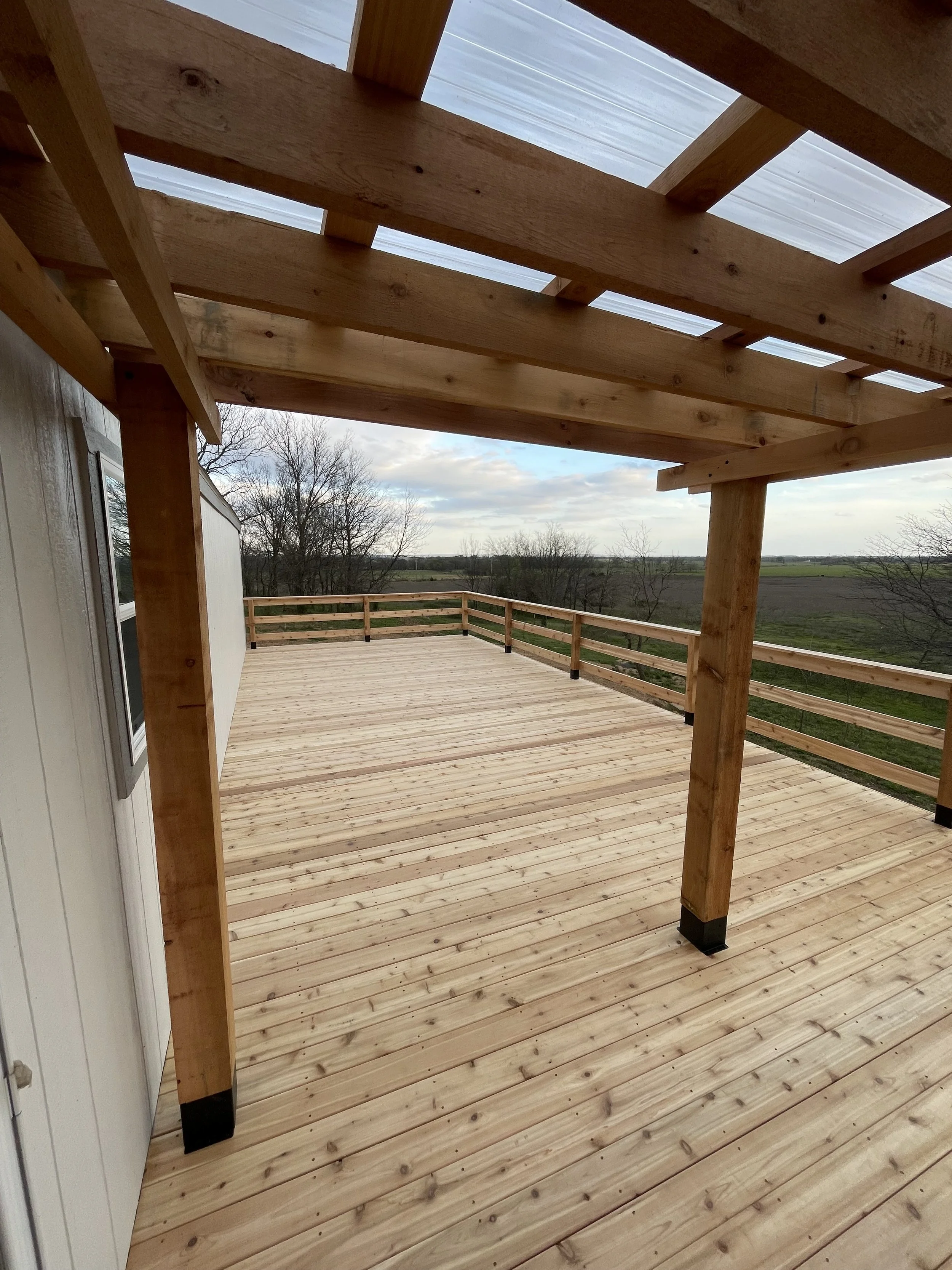 Wooden deck with a partially constructed pergola, overlooking a rural landscape with trees and open land.