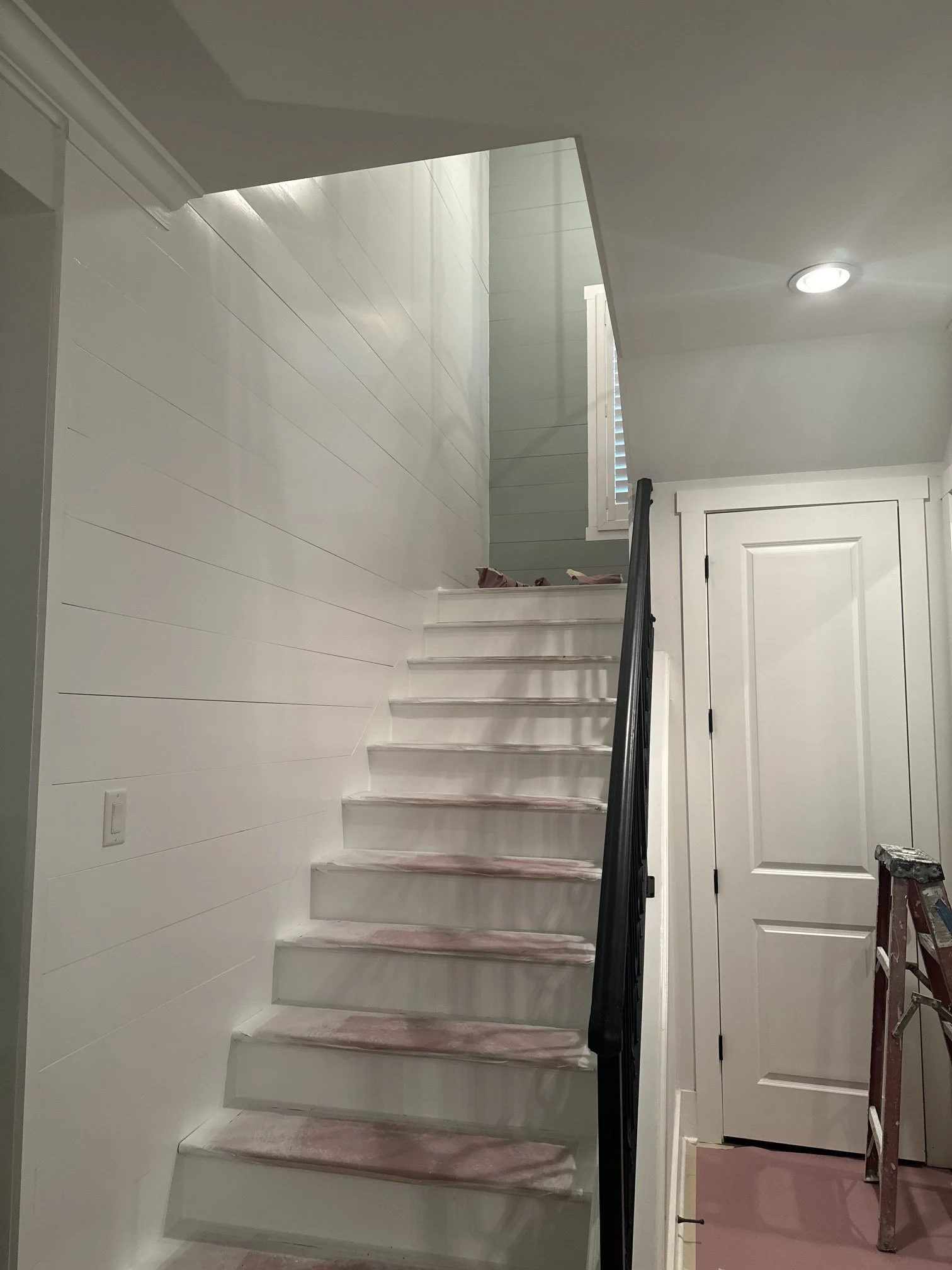 Interior of a house under renovation showing a staircase with steps covered in pink protective paper, a white wall with horizontal paneling, a window with white shutters, a closed white door, and a small ladder.