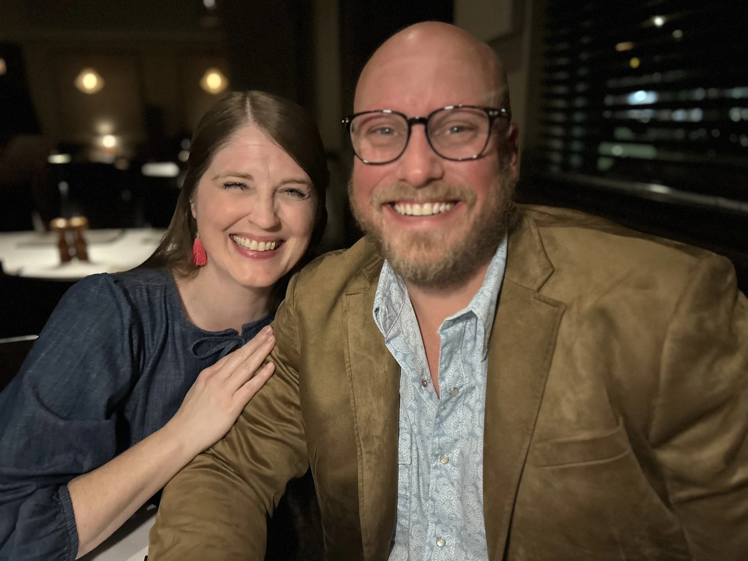 Smiling woman with brown hair and red earrings, and smiling man with glasses and a beard, sitting close together in a dimly lit restaurant.
