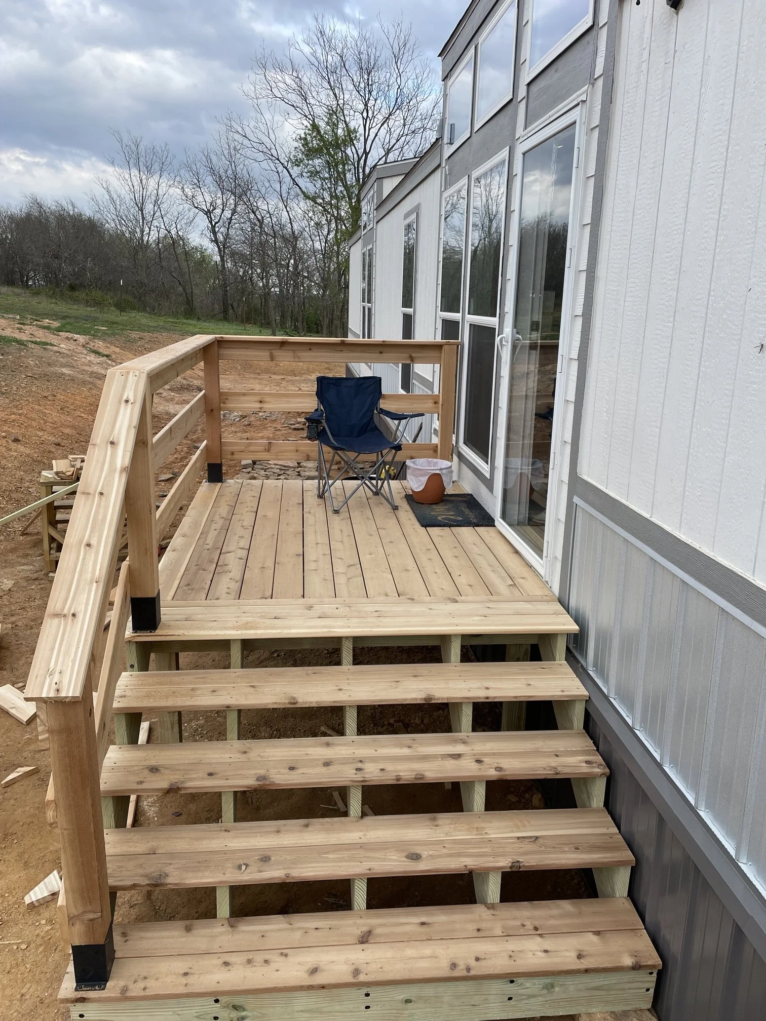 A partially constructed wooden deck outside a house with large windows and a glass sliding door, with a folding chair and a small pot on the deck, surrounded by a natural landscape with trees and cloudy sky.