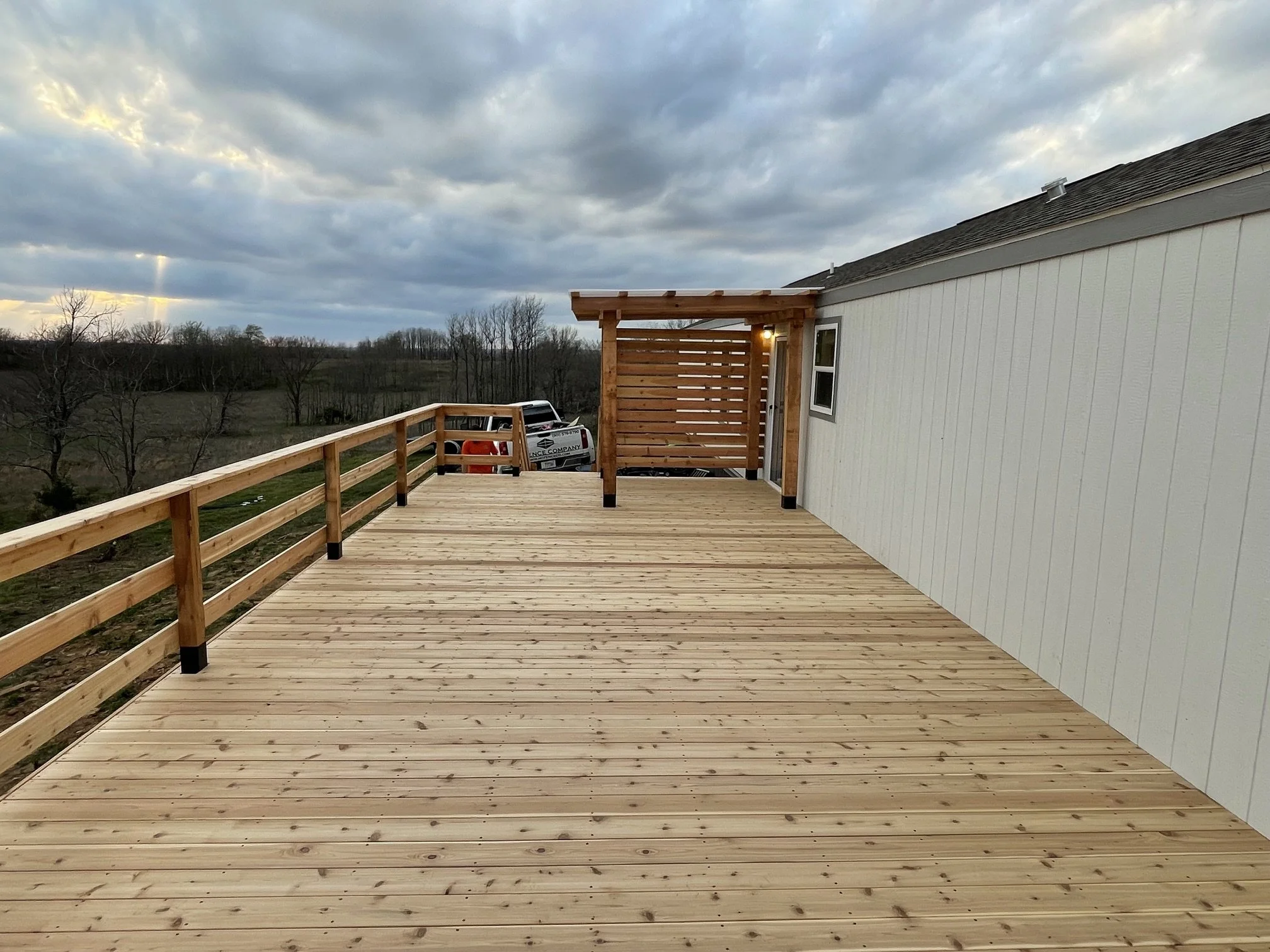 A wooden deck attached to a house with a railing and a privacy screen, overlooking a rural landscape with leafless trees and a cloudy sky.