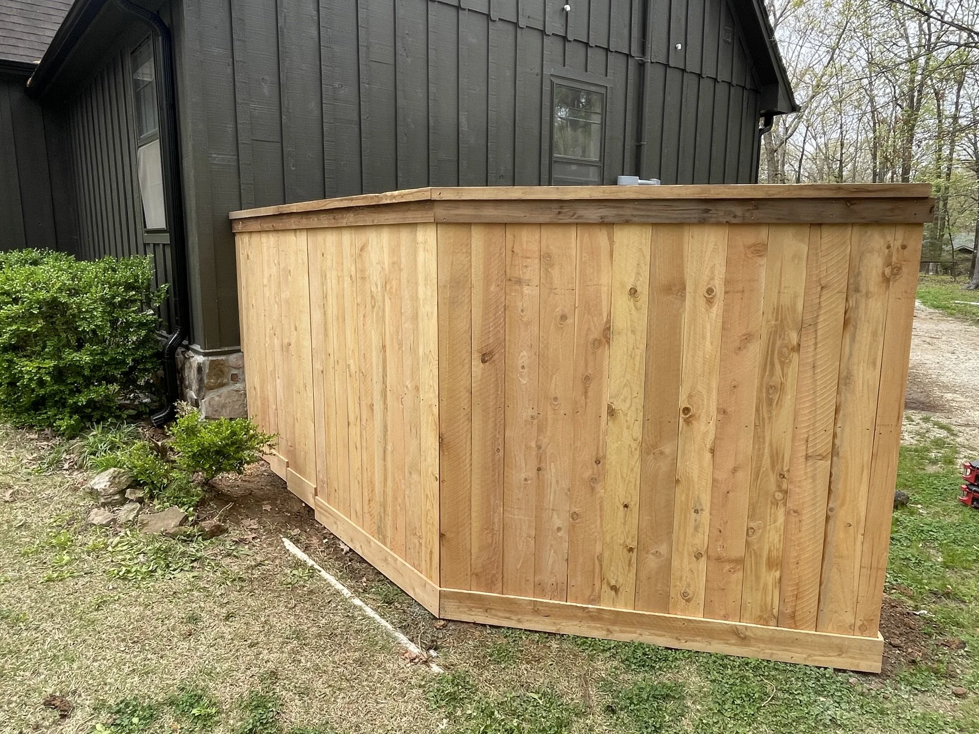 A newly built curved wooden privacy fence against the side of a dark-colored house with a lawn and trees in the background.
