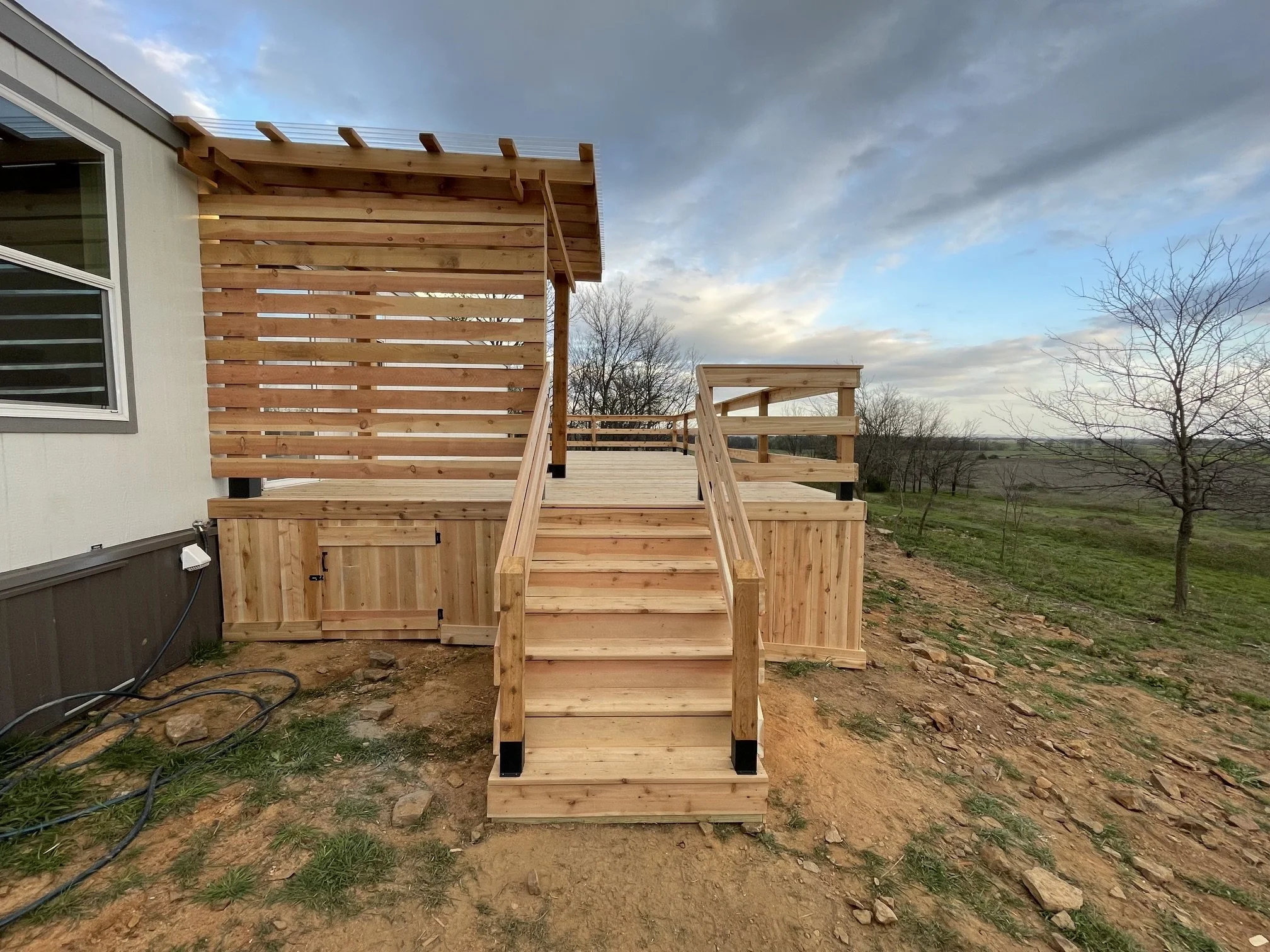 Unfinished wooden deck with stairs extending from the side of a house, with a wooden privacy fence and a railing. Overcast sky with dark clouds, trees and a field in the background.