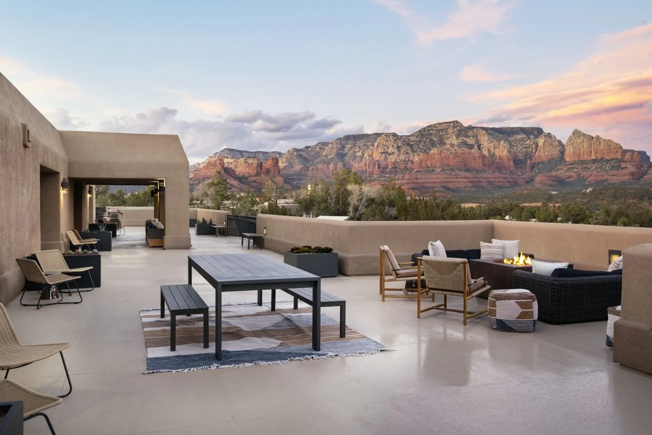 Outdoor rooftop patio with beige walls, modern outdoor furniture, a fire pit, and scenic red rock mountains in the background during sunset.