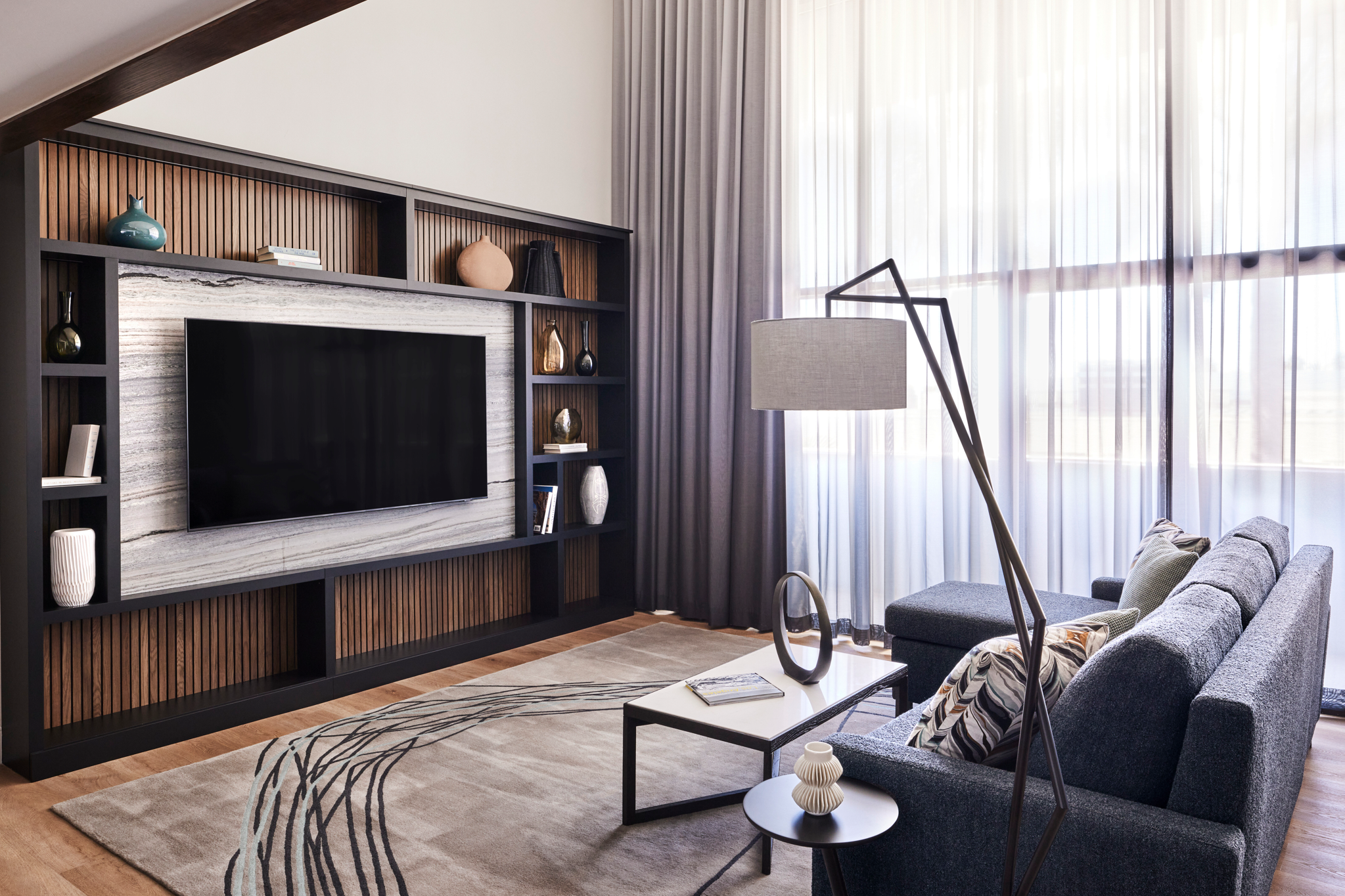 Modern living room with a gray sofa, patterned pillows, a small black coffee table, and a beige and black rug. There is a large window with sheer white curtains, a tall black floor lamp, and a black shelving unit with decorative vases and books. The 