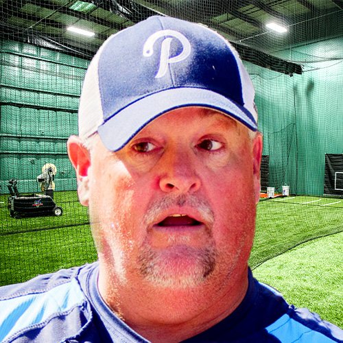 Man in a blue and white baseball cap and athletic shirt standing in an indoor sports facility with green turf and batting cages.