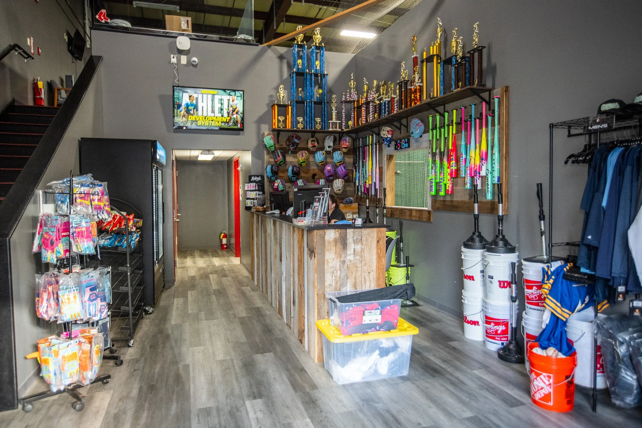 A sports store interior with a display of baseball bats, helmets, and gear. It features a wooden counter, a wall of trophies, and shelves stocked with merchandise.