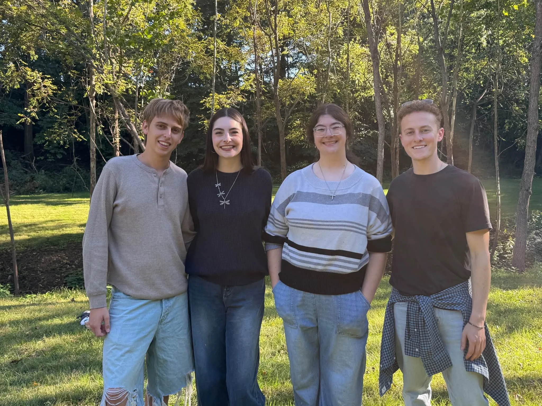 Four young adults standing outdoors on a grassy area with trees and sunlight in the background, smiling and looking at the camera.