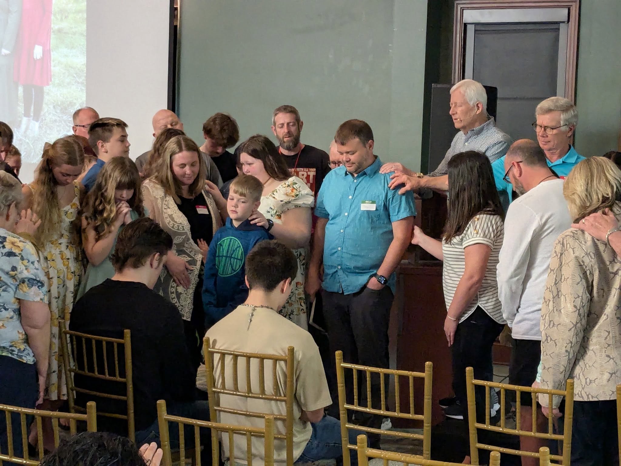 A large group of people, including children and adults, are gathered in prayer or reflection, with some adults placing their hands on children's shoulders, in a room with a green wall and gold chairs.