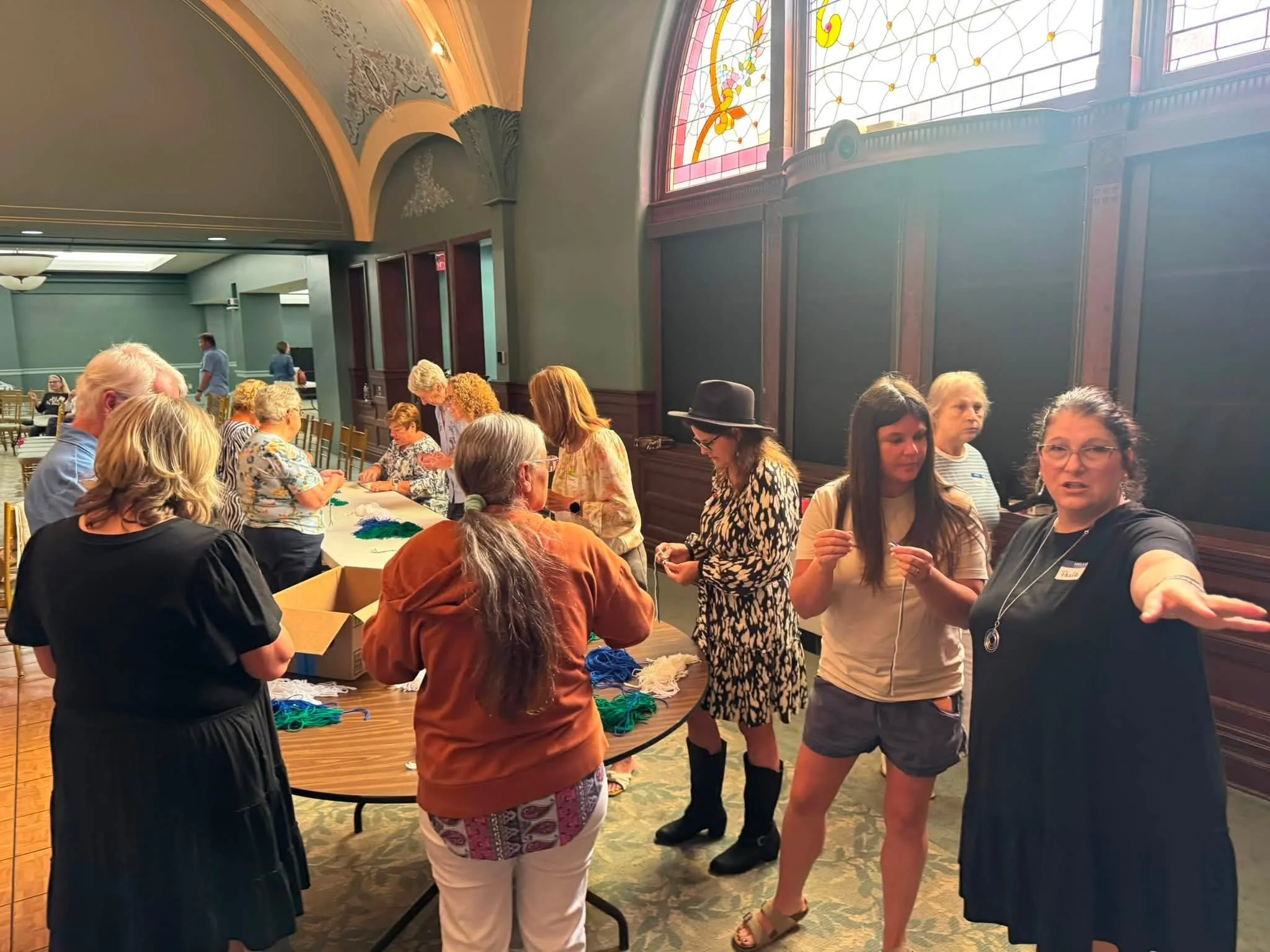 People gathered in a room with stained glass windows and ornate architectural details, participating in a craft or social event at a table with colorful yarn.