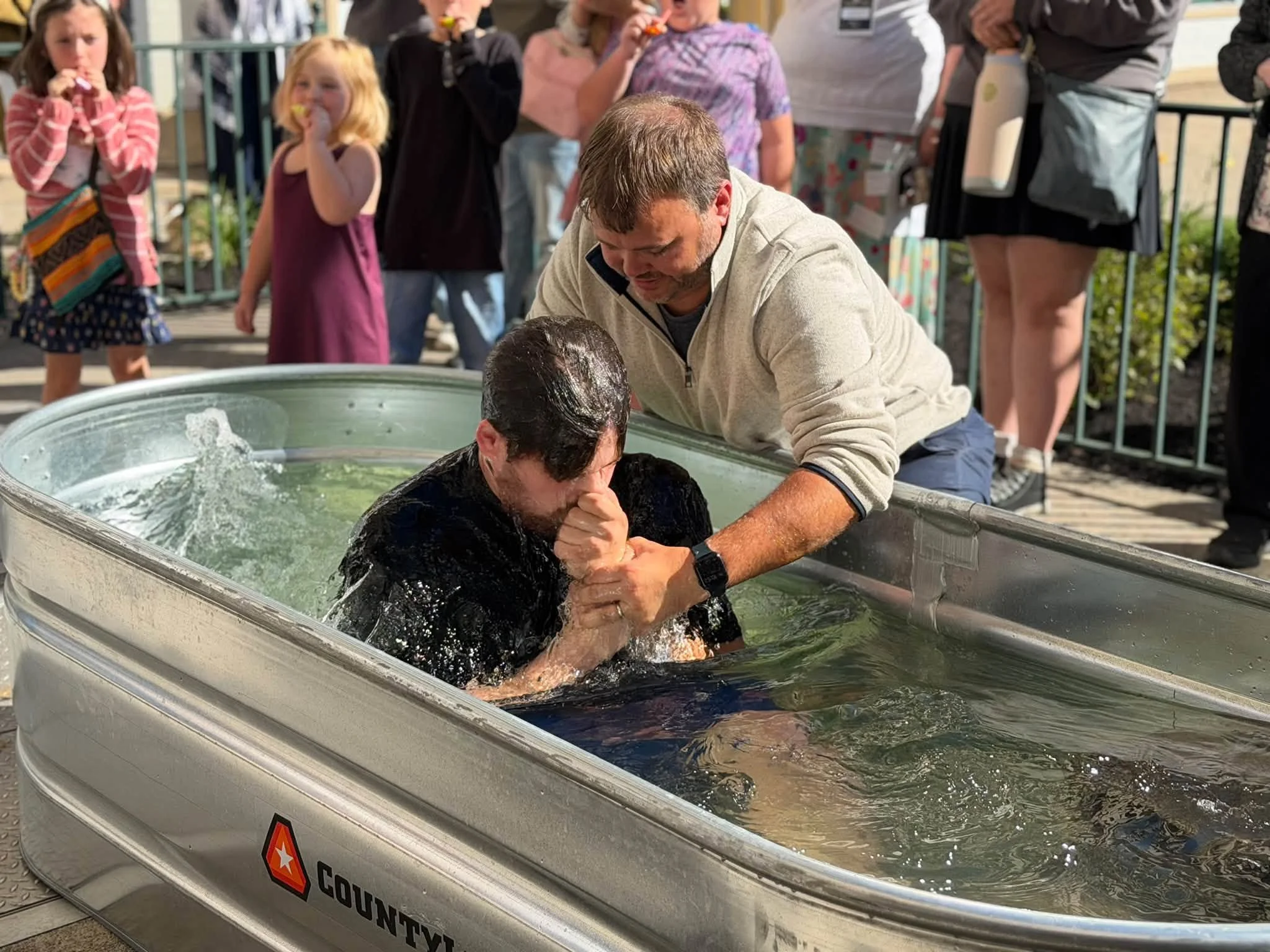 A man helping a person baptize another person in a metal tub filled with water, with a group of onlookers including children watching nearby.