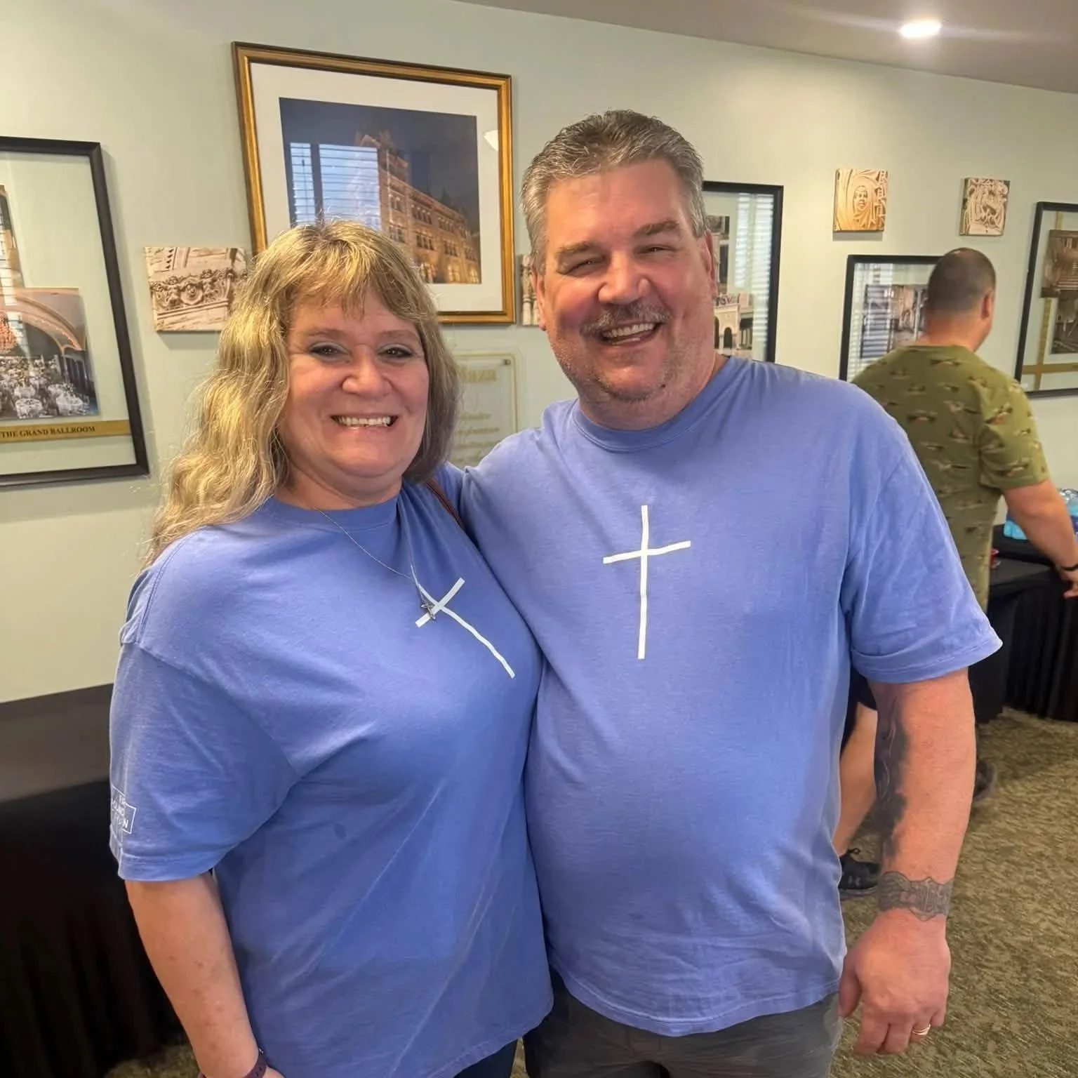 A smiling woman and man wearing matching blue T-shirts with white crosses, standing indoors with framed pictures on the wall behind them.