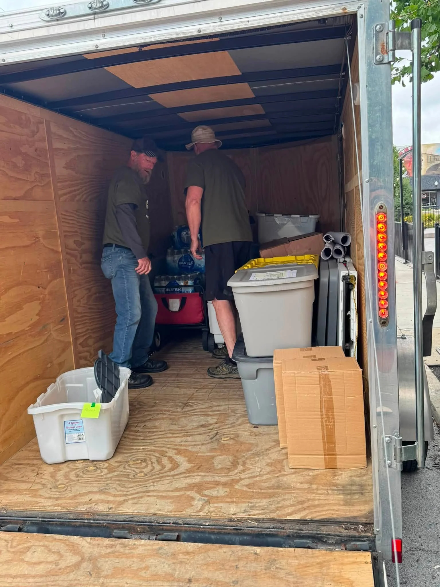 Two men inside an empty wooden cargo trailer, surrounded by various storage containers and rolled-up posters or blueprints, with an outdoor scene visible on the right side.
