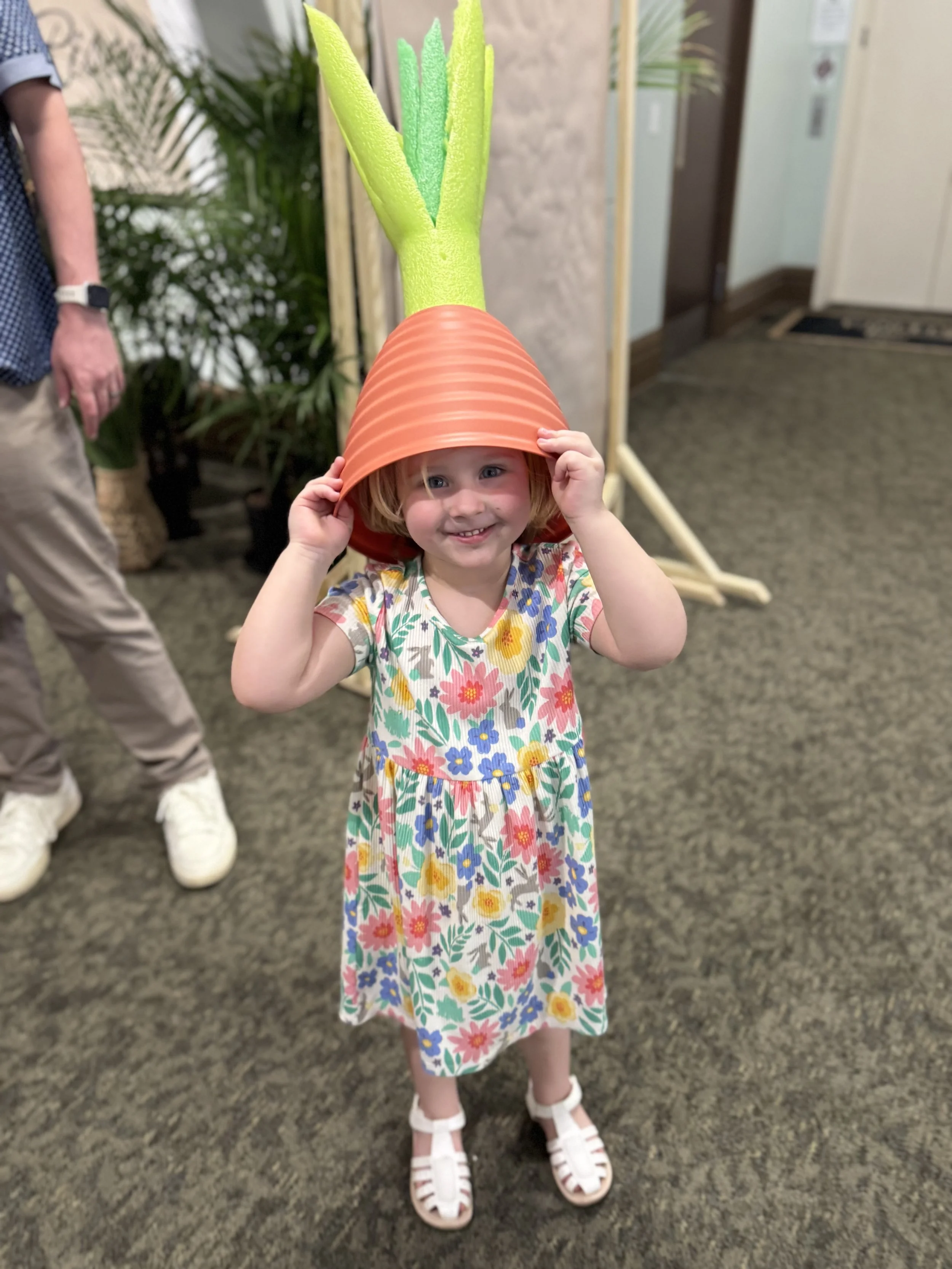 A young girl wearing a floral dress and white sandals holds a terracotta pot upside down on her head, with a green foam plant decoration placed on top, in an indoor setting with carpeted flooring and potted plants in the background.