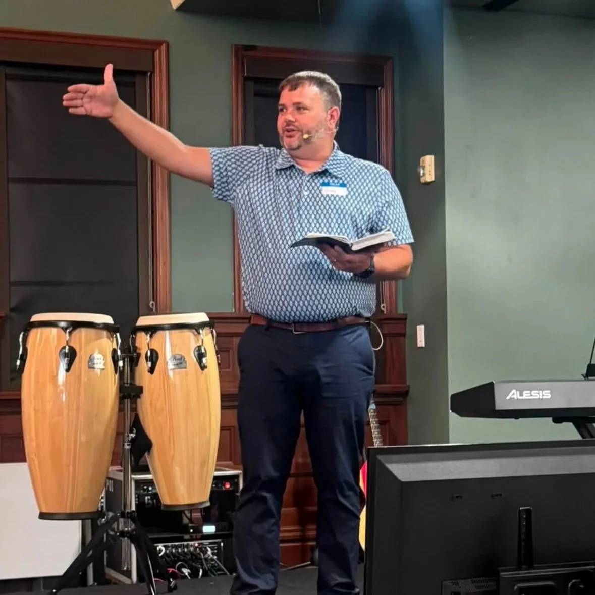 A man giving a speech in a room with musical instruments, including conga drums and a keyboard, while holding a book.