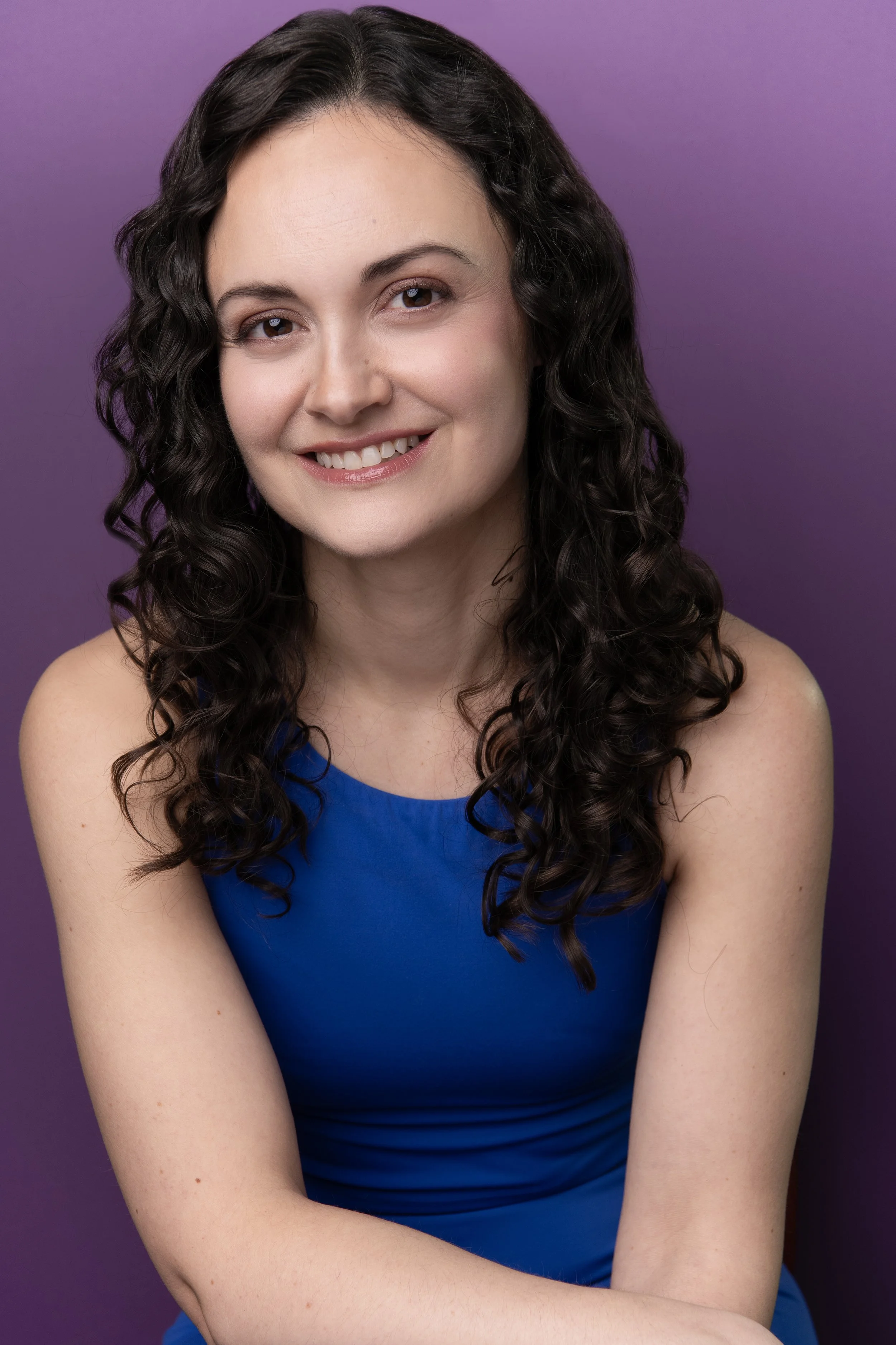 A woman with long, curly brown hair, smiling while wearing a black sleeveless top, in an indoor setting with warm lighting.