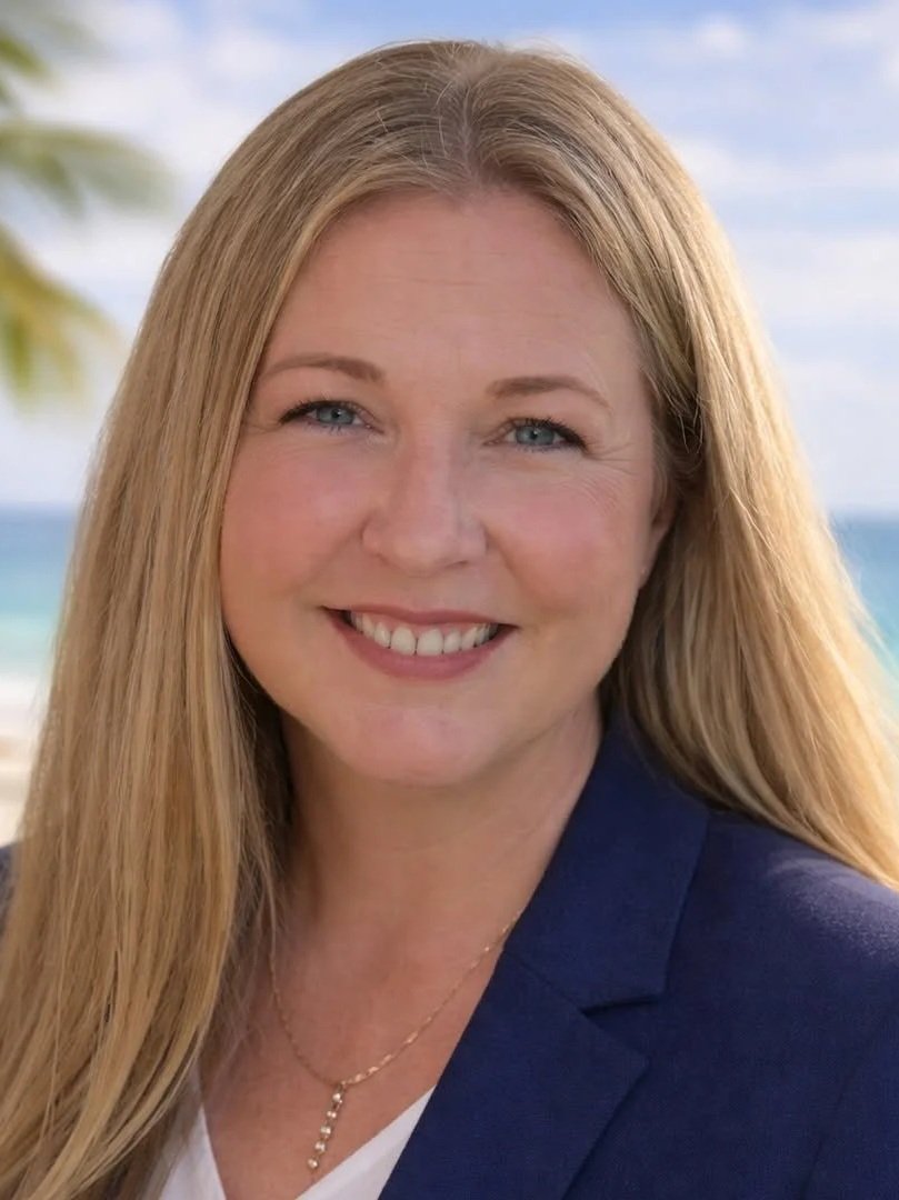 Close-up portrait of a woman with long blonde hair, blue eyes, smiling, wearing a navy blazer and a delicate necklace, outdoors near the beach with palm trees and ocean in the background.