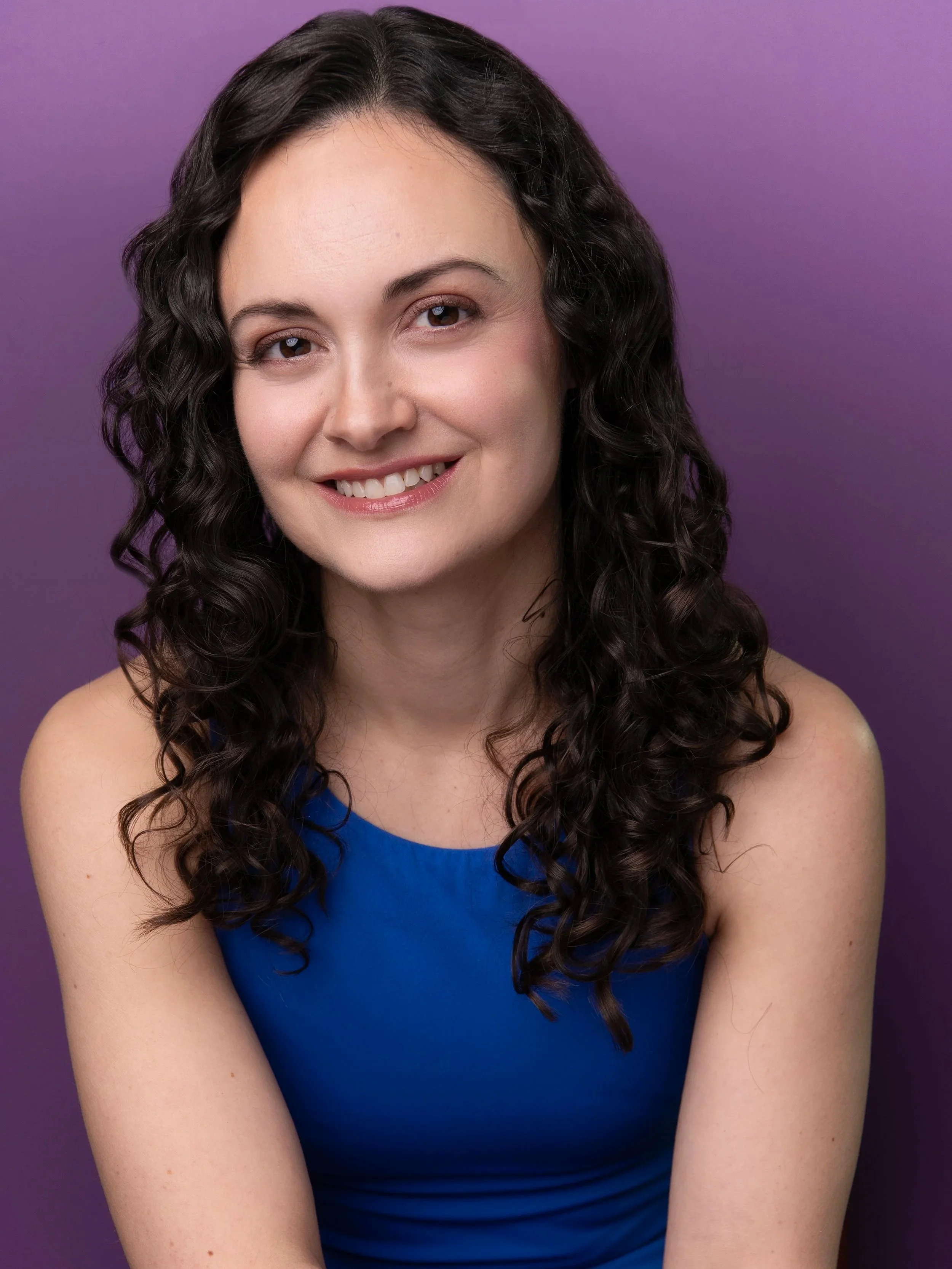 Close-up portrait of a young woman with long, curly dark hair, smiling, wearing a blue top, against a purple background.
