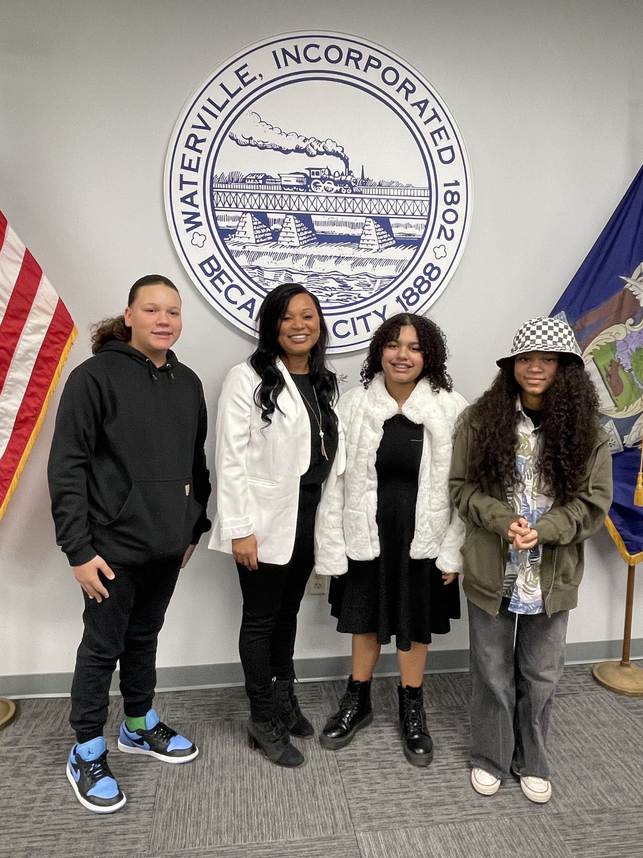 Flavia DeBrito with three young people in front of the City of Waterville seal
