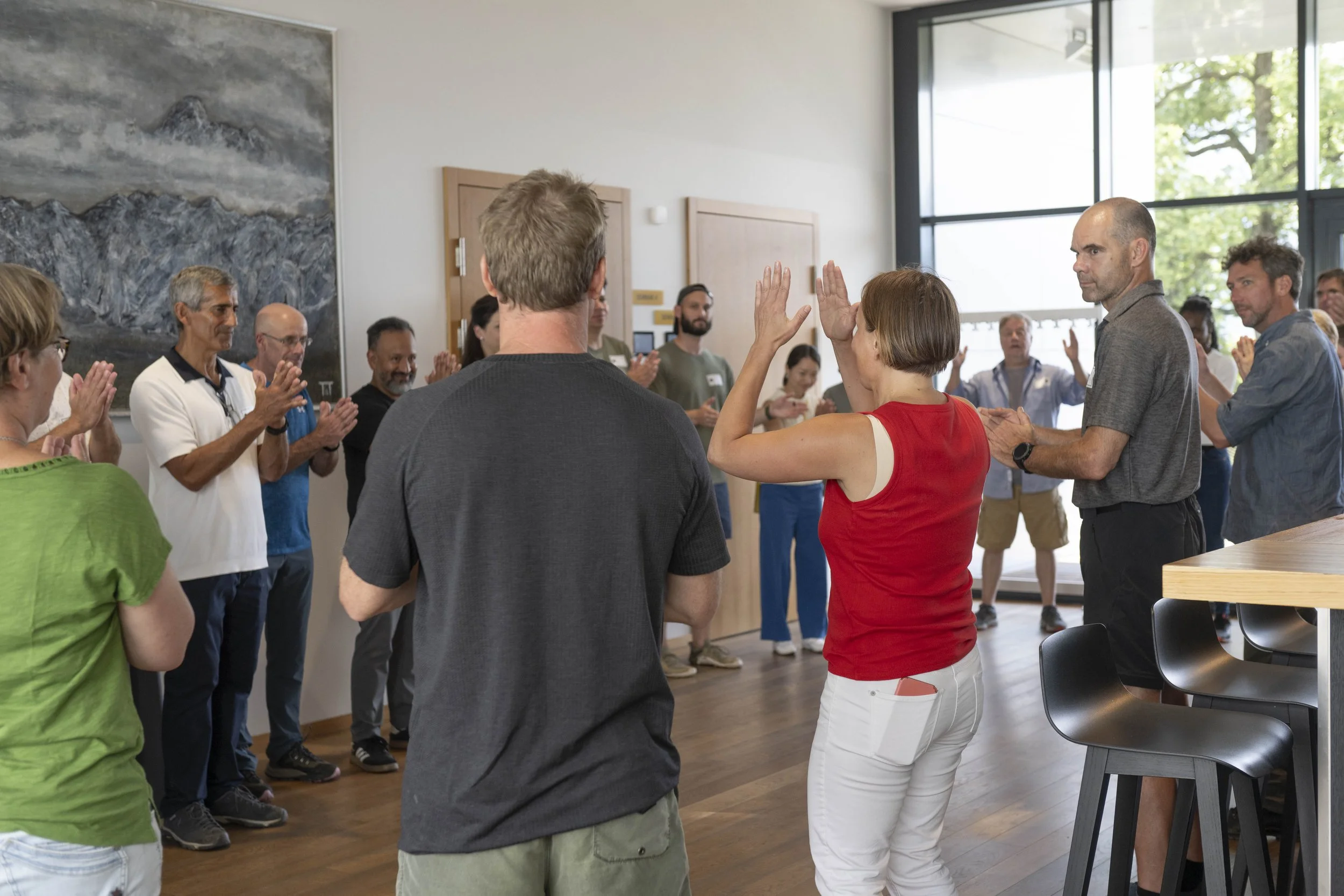 A diverse group of people in a circle participating in an experiential workshop in a modern room with large windows and trees outside.