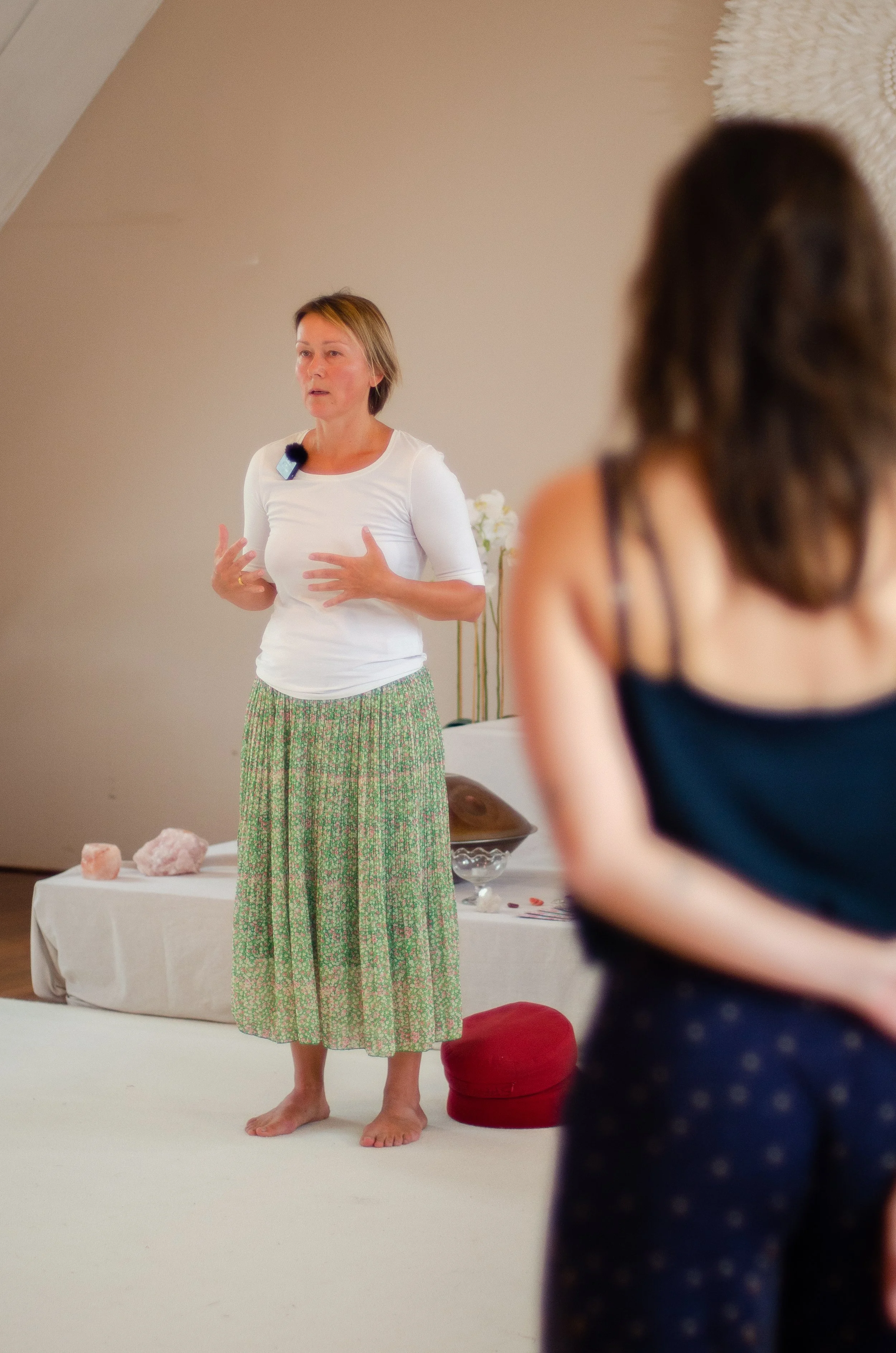 A woman standing barefoot on a white floor, speaking to an audience, with a blurred person in the foreground. The woman has short hair, is wearing a white shirt and green patterned skirt, and is gesturing with her hands while talking. Behind her, there is a table with rocks and flowers, and a beige wall.