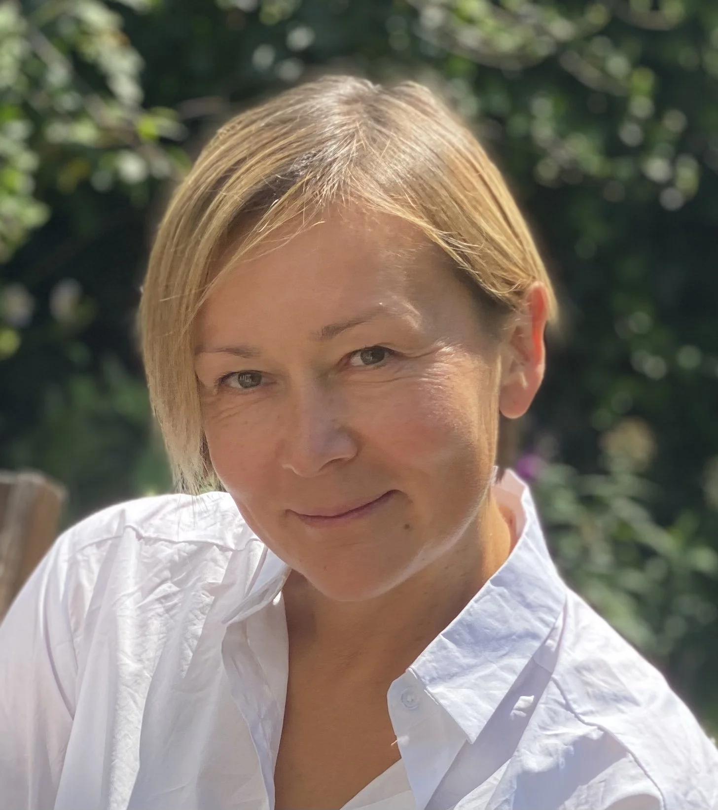 Close-up of a woman with short blonde hair wearing a white shirt, outdoors with green foliage in the background.
