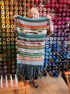 A woman holding a colorful striped woven blanket in a yarn shop with shelves of multicolored yarn behind her.