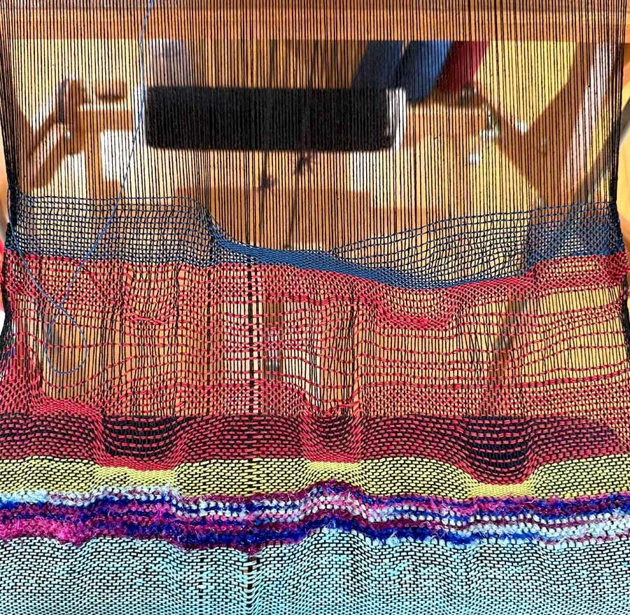 Colorful woven textile fabric with intricate patterns and a mesh curtain in the foreground, wooden surface in the background, partially visible objects, and a person mirrored in the reflection.