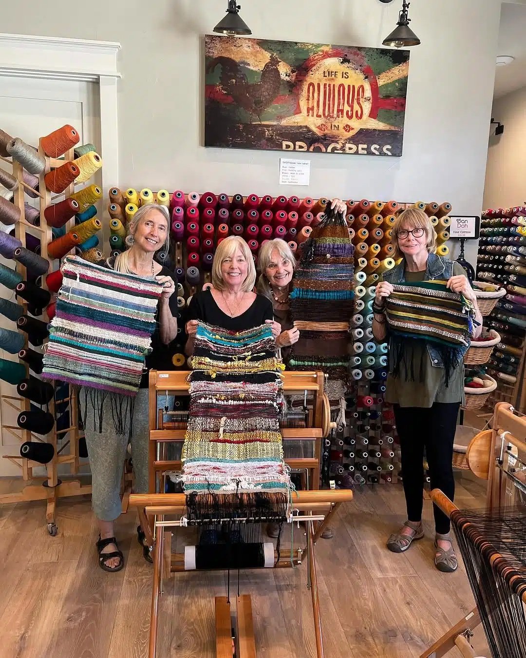 Four women standing in a yarn and weaving store, holding colorful woven textiles. Behind them are shelves filled with colorful spools of thread and yarn. They are smiling and appear to be proud of their work.